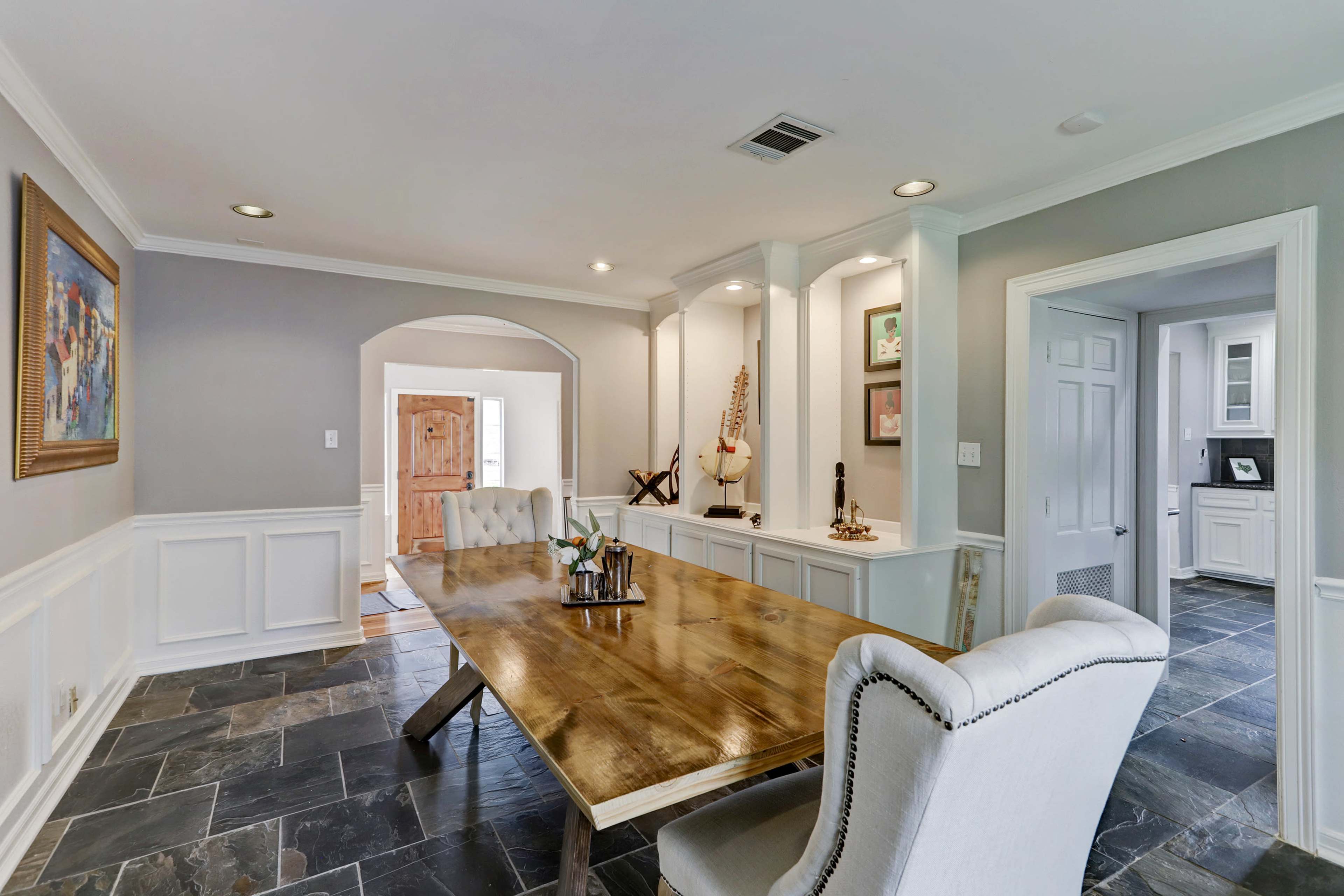 A wooden dining table with decorative items is centered in a well-lit room, framed by white wainscoting and gray walls, with doors leading to adjacent spaces.