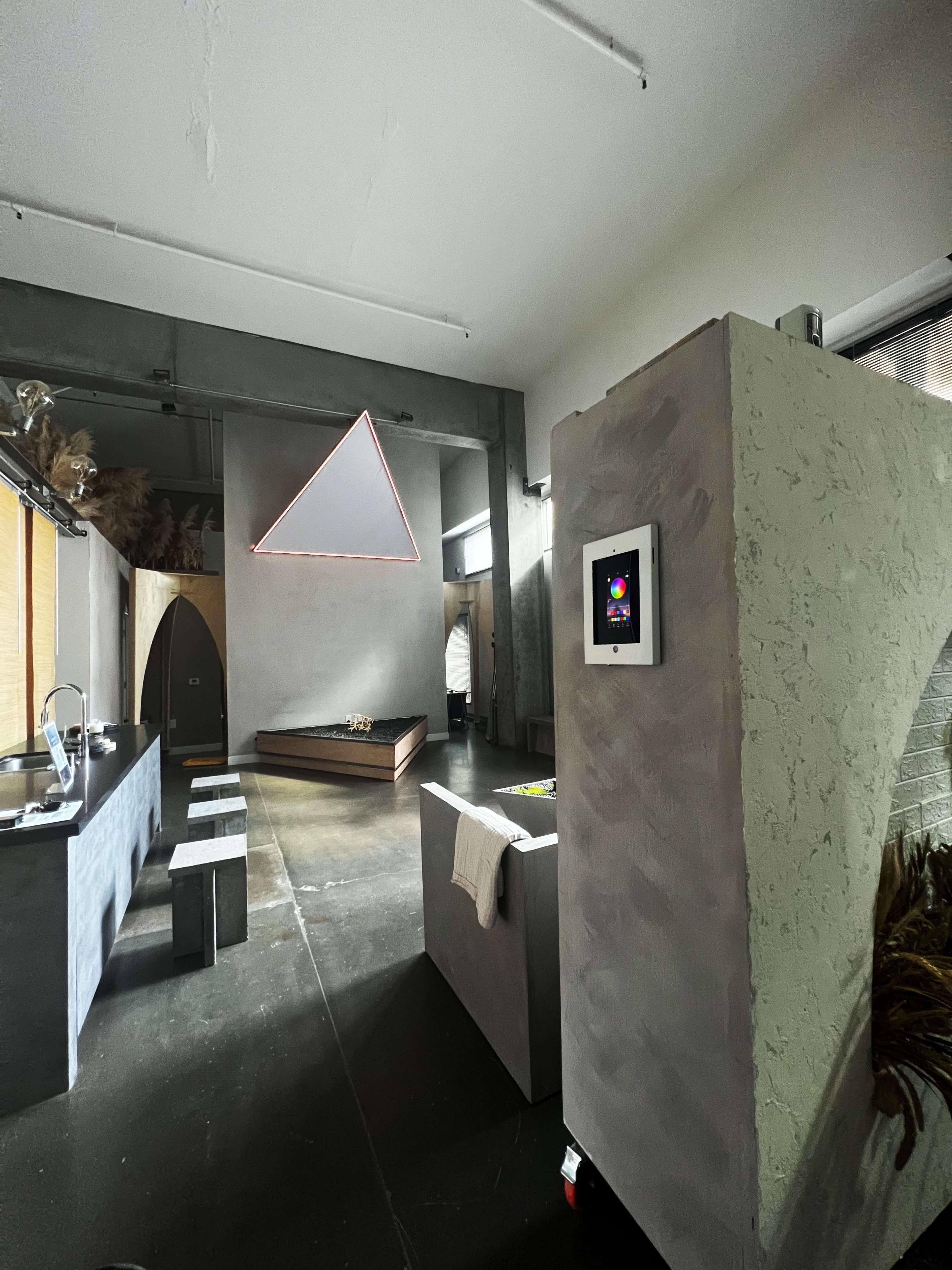 The image shows a modern interior space with an open layout, featuring a kitchen area, a minimalist bed, and a geometric light fixture hanging from the ceiling.