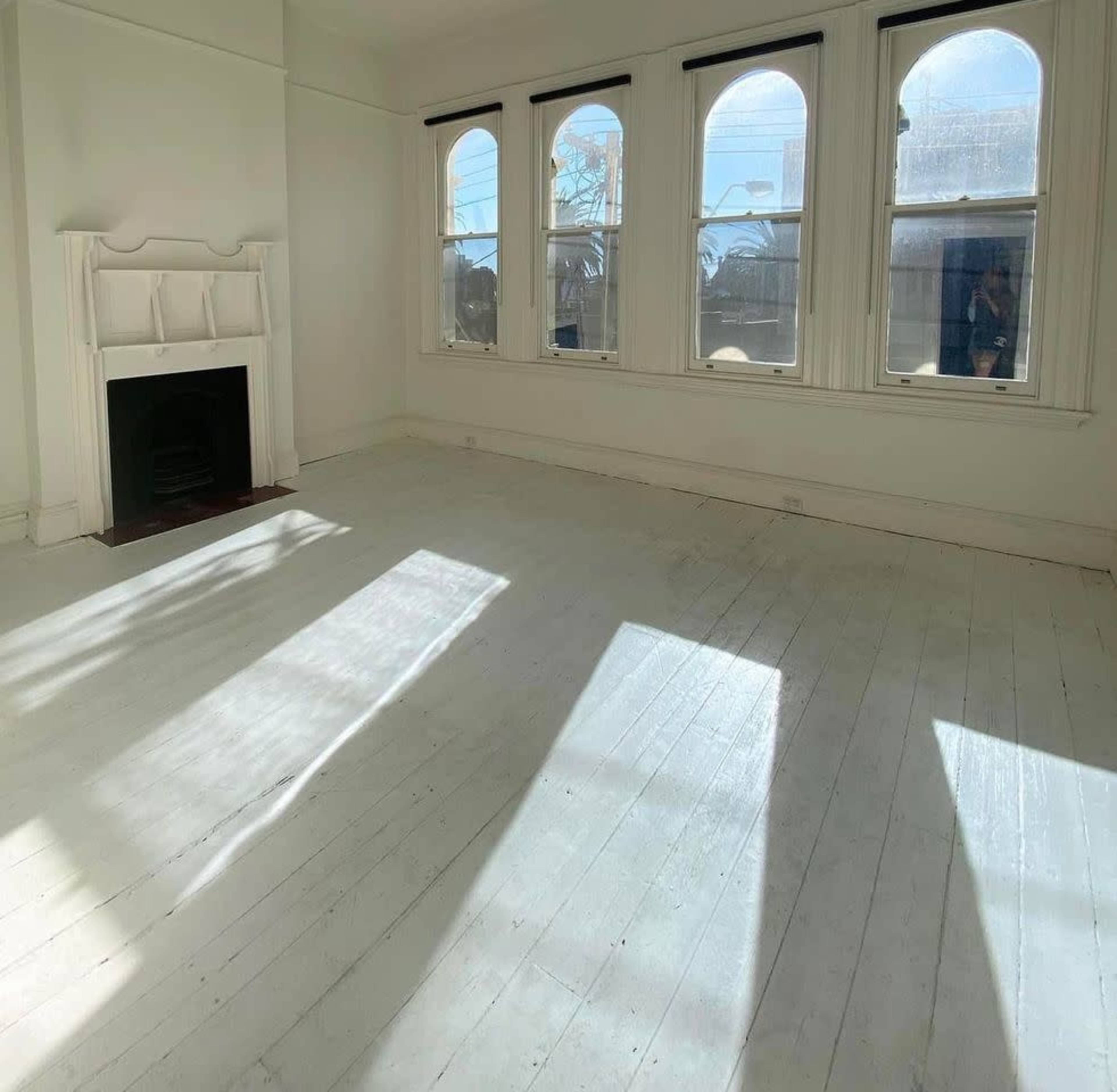 The image shows a bright, empty room with large windows and a fireplace, featuring wooden floors that reflect light.