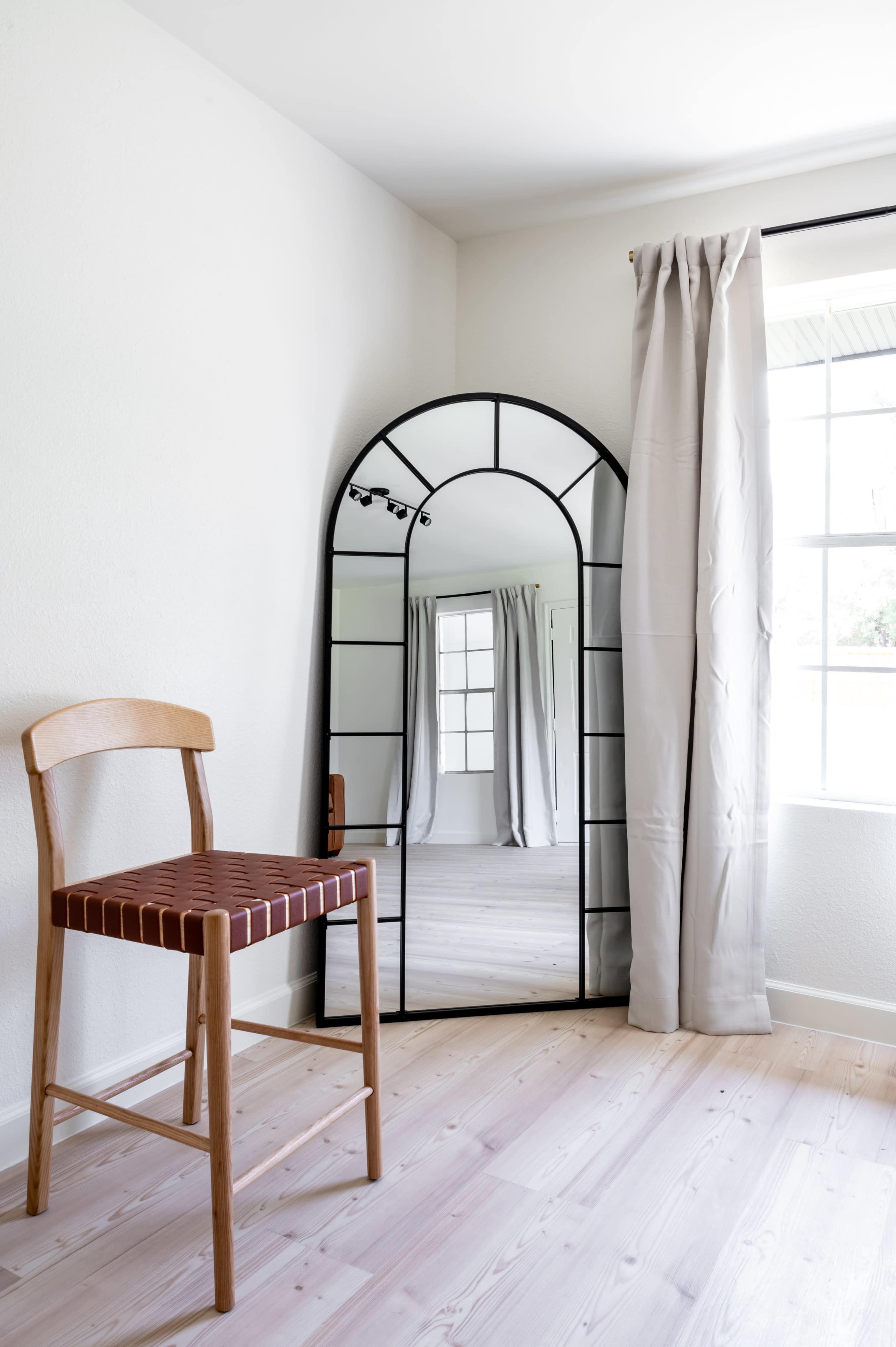 A wooden chair with a striped seat is positioned next to a large, arched mirror reflecting a bright room with a window and long curtains.