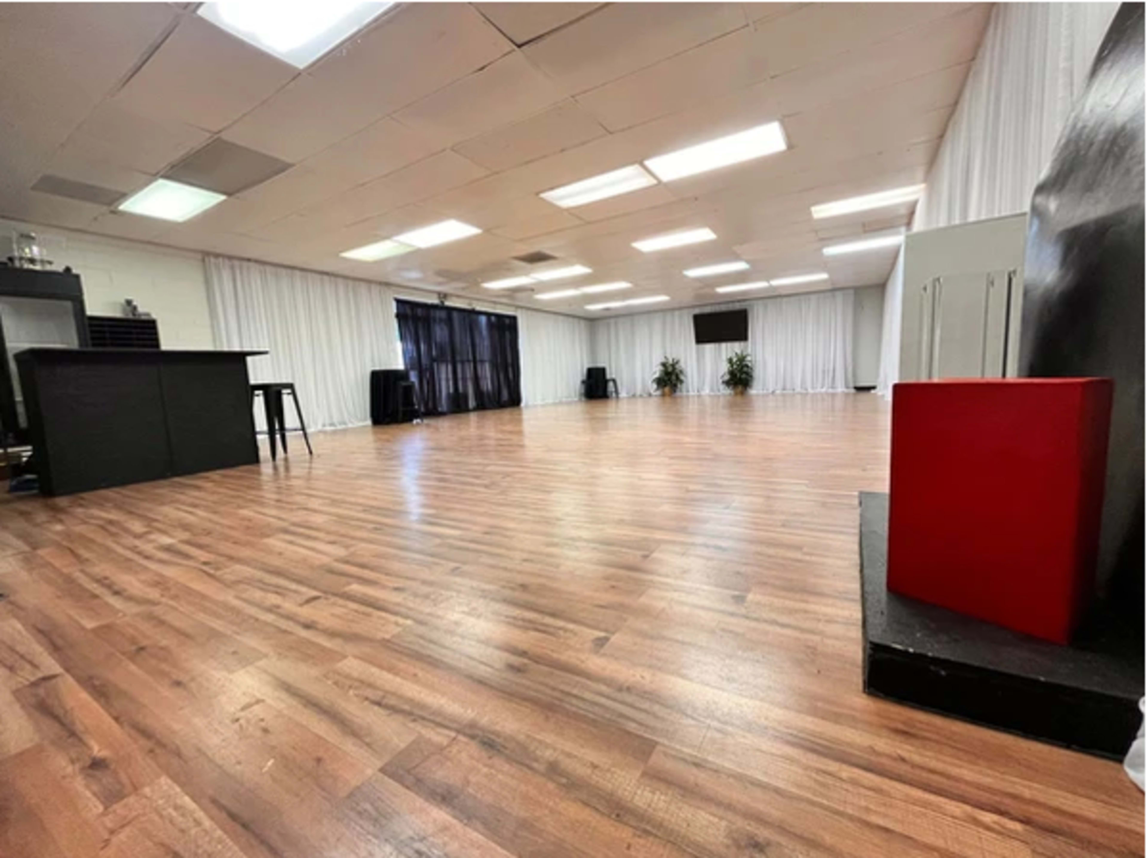 A spacious, well-lit room with wooden flooring, white curtains, and minimalist black furniture.