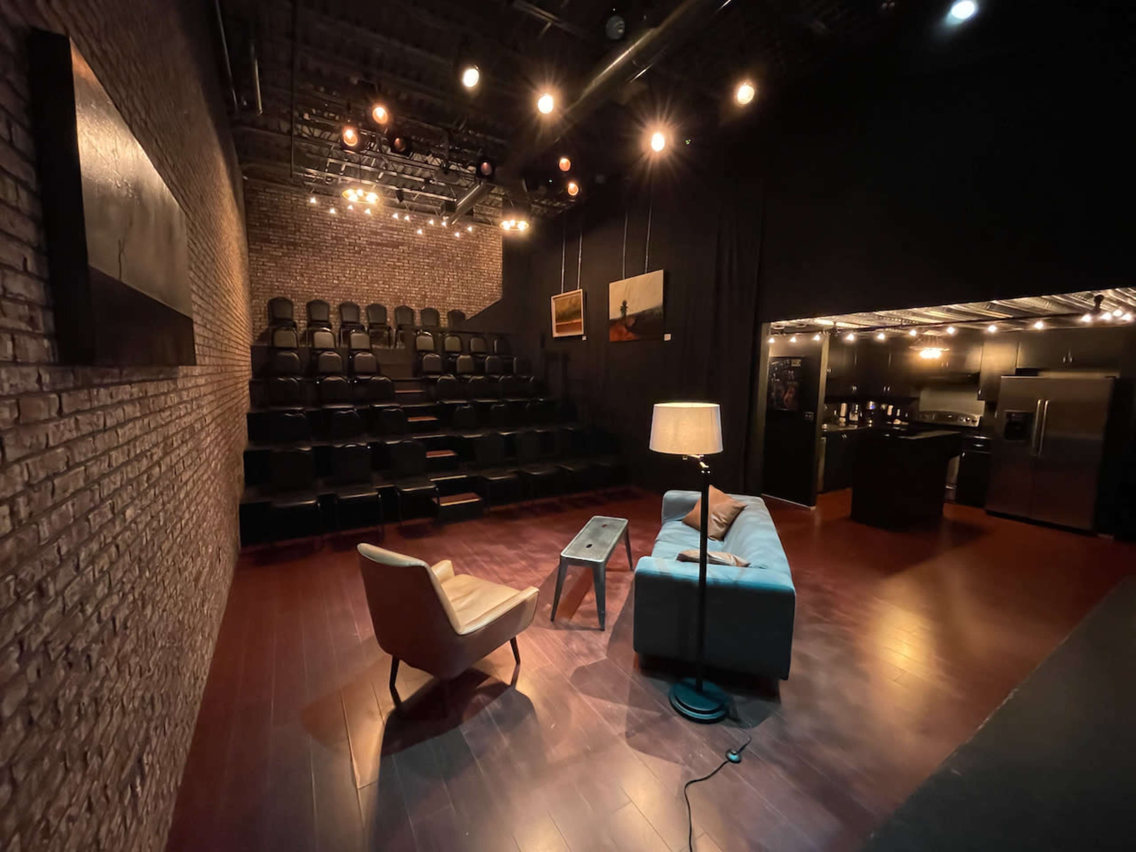 The image shows a small theater setting with tiered seating, a gray sofa, a lamp, and a kitchenette area in the background.