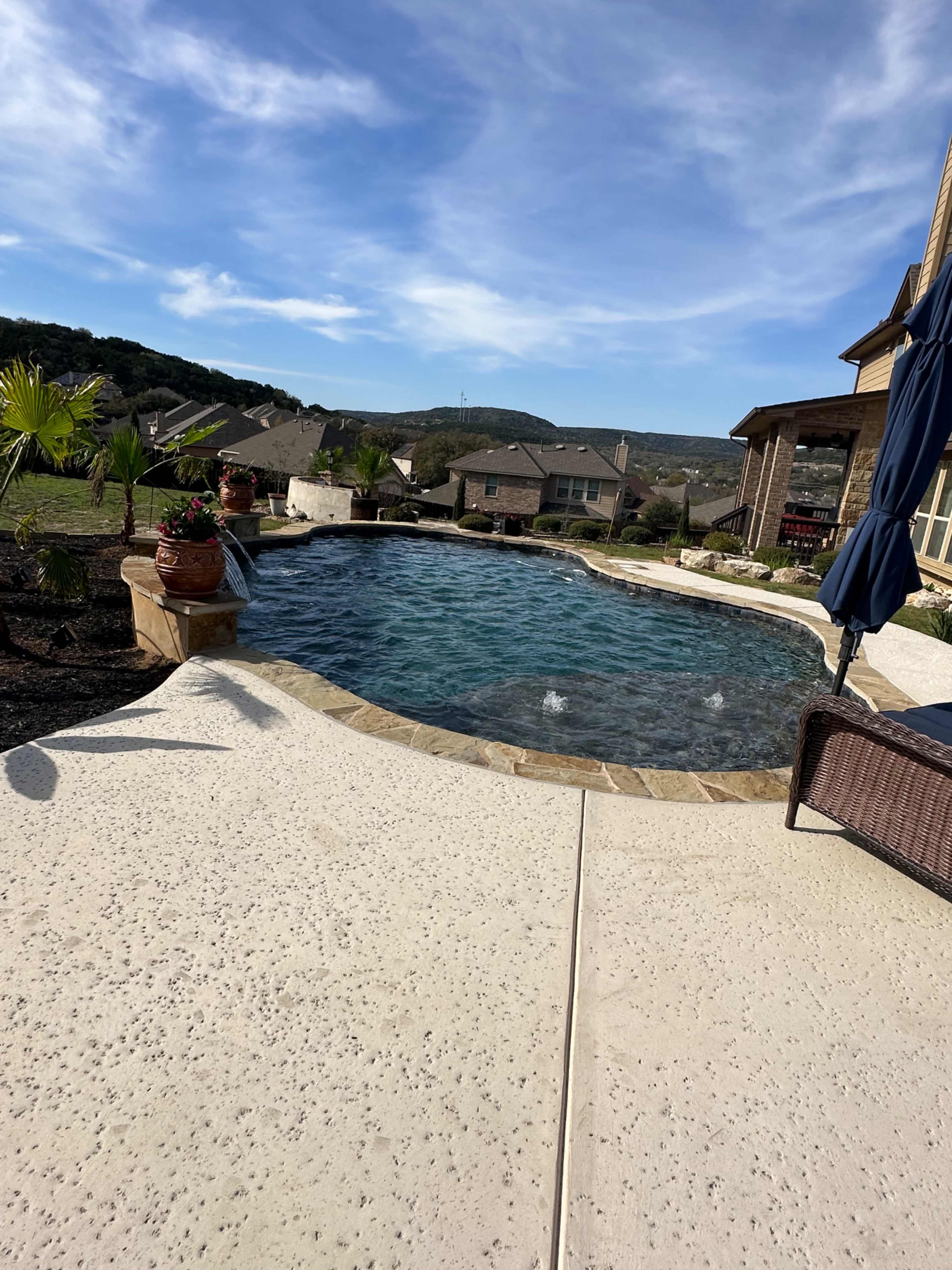 The image shows a swimming pool surrounded by potted plants and a patio area, with houses and hills in the background under a blue sky.