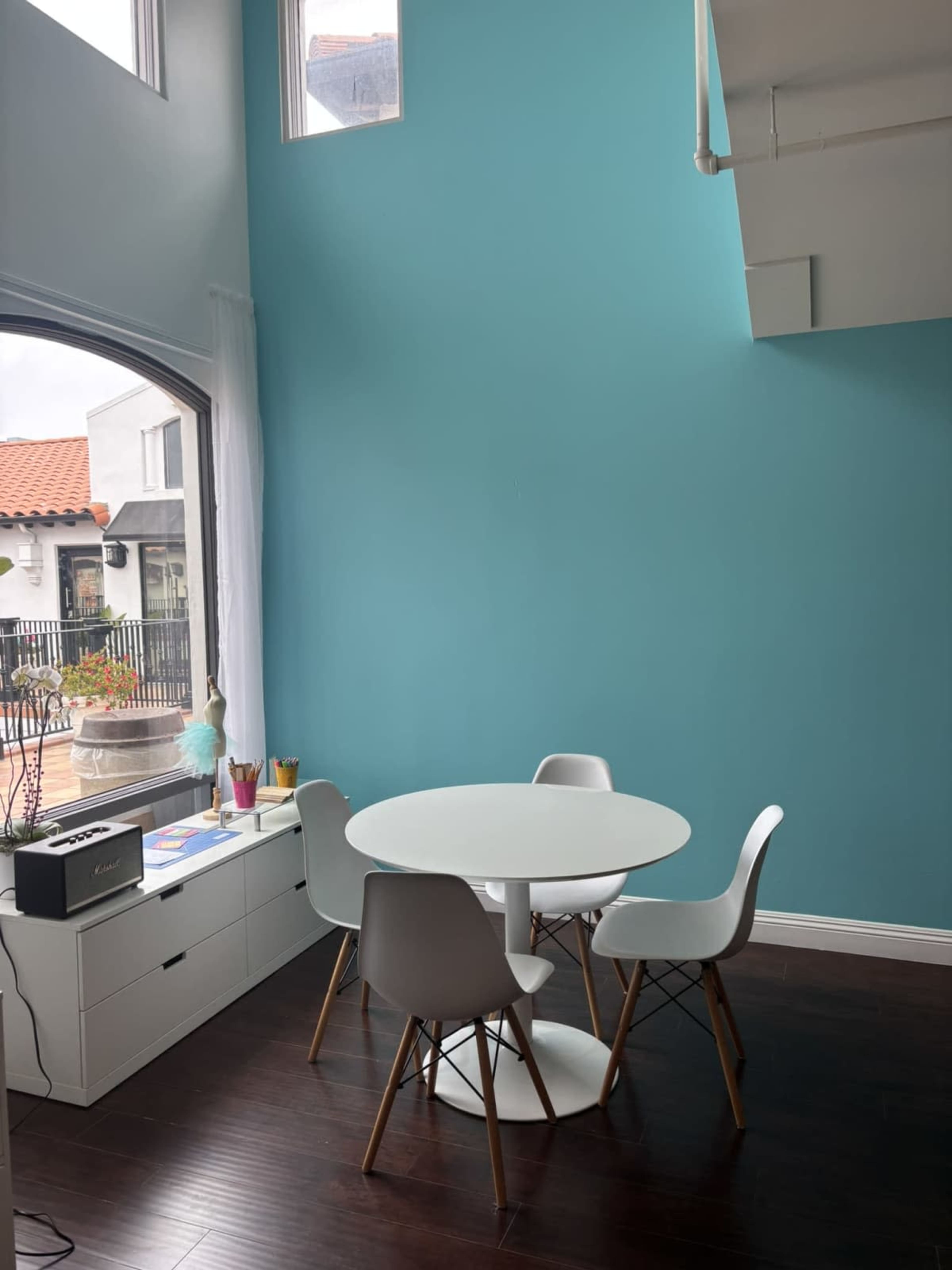 The image shows a small dining area with a round white table and four white chairs against a bright blue wall, featuring a window and a dresser with various items on it.
