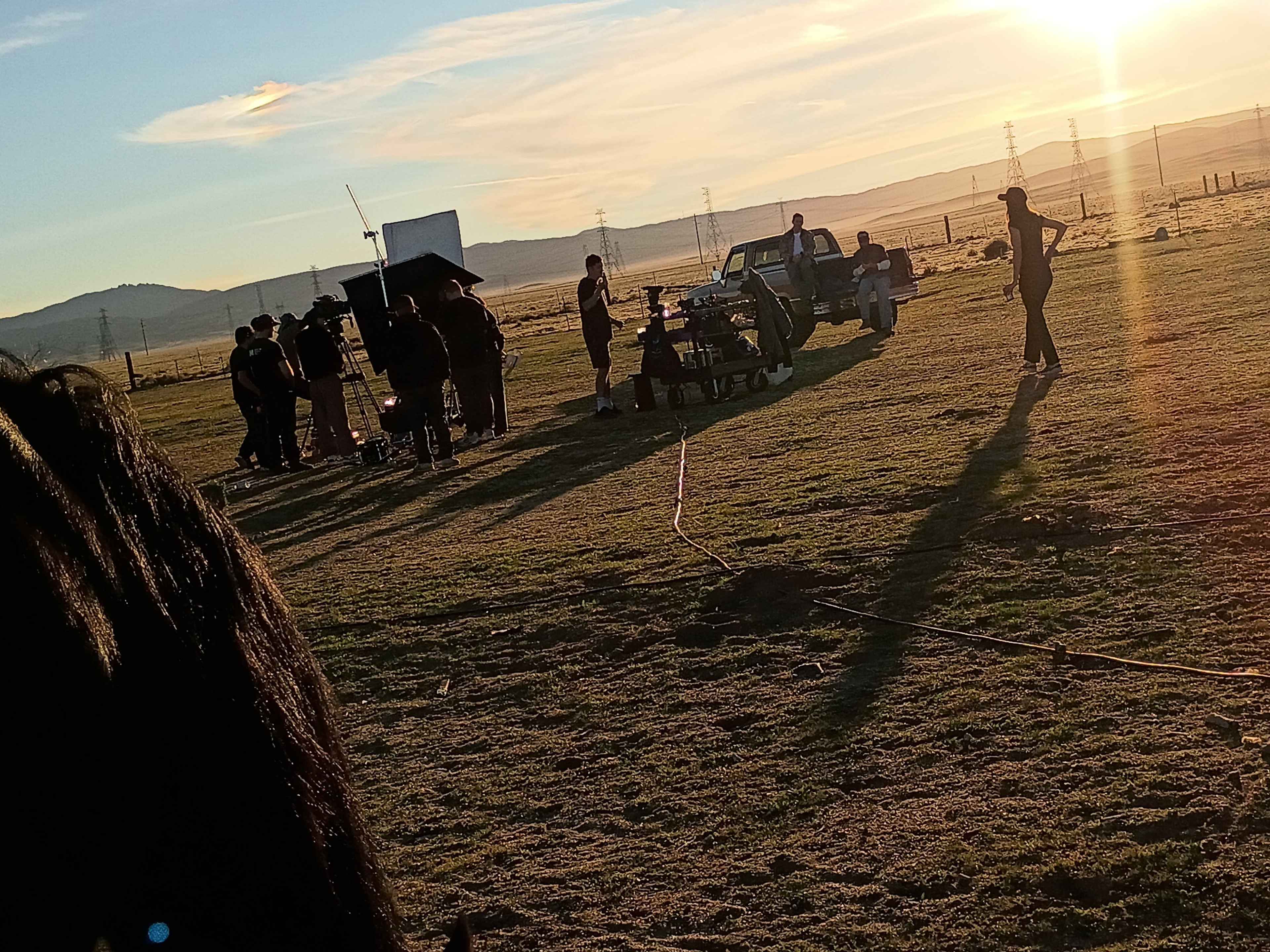A film crew is set up outdoors near a vehicle, filming during sunset with multiple people and equipment scattered across a grassy field.