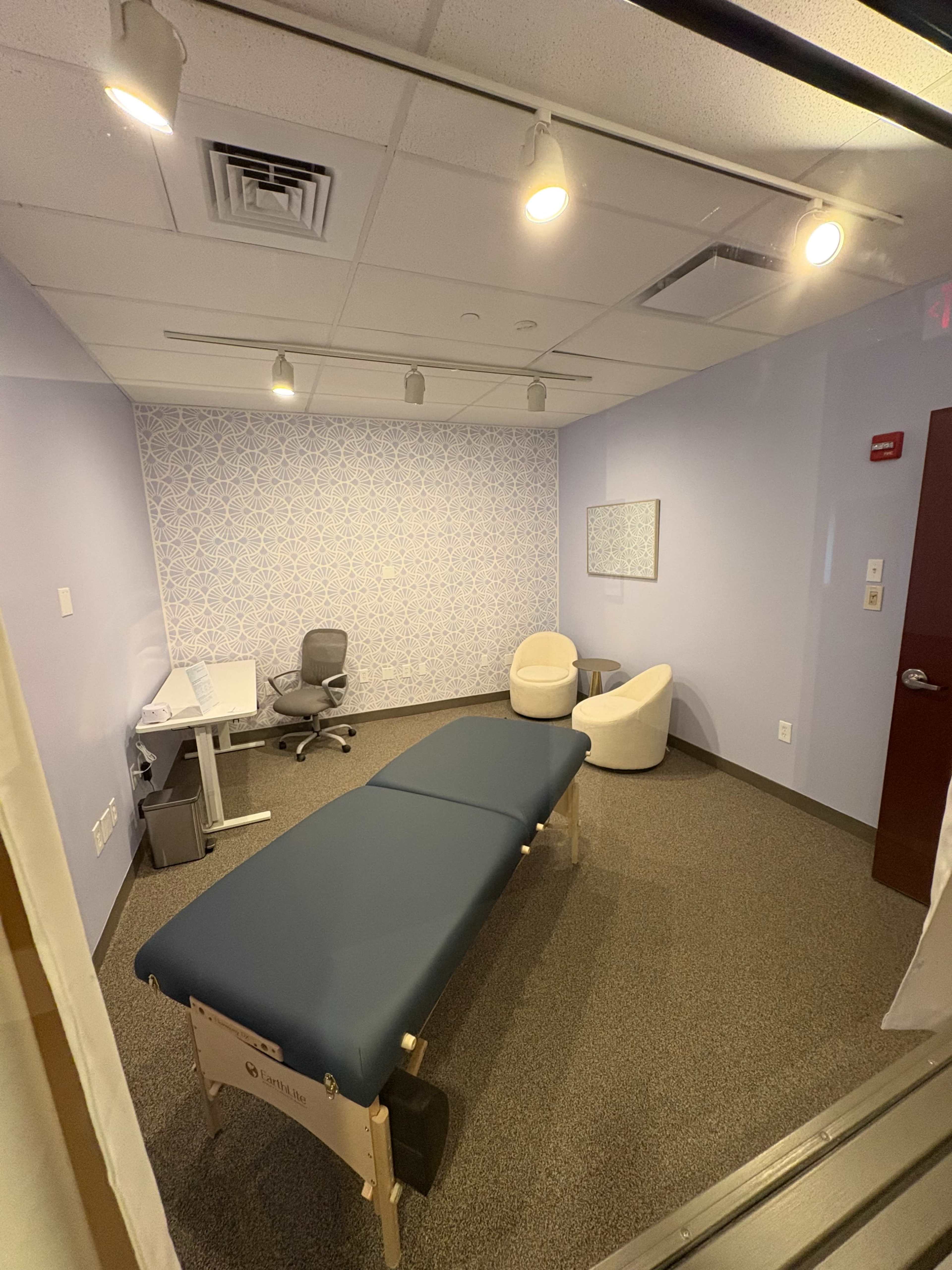 The image shows a small treatment room with a massage table, two chairs, a desk, and blue walls adorned with a patterned accent wall.