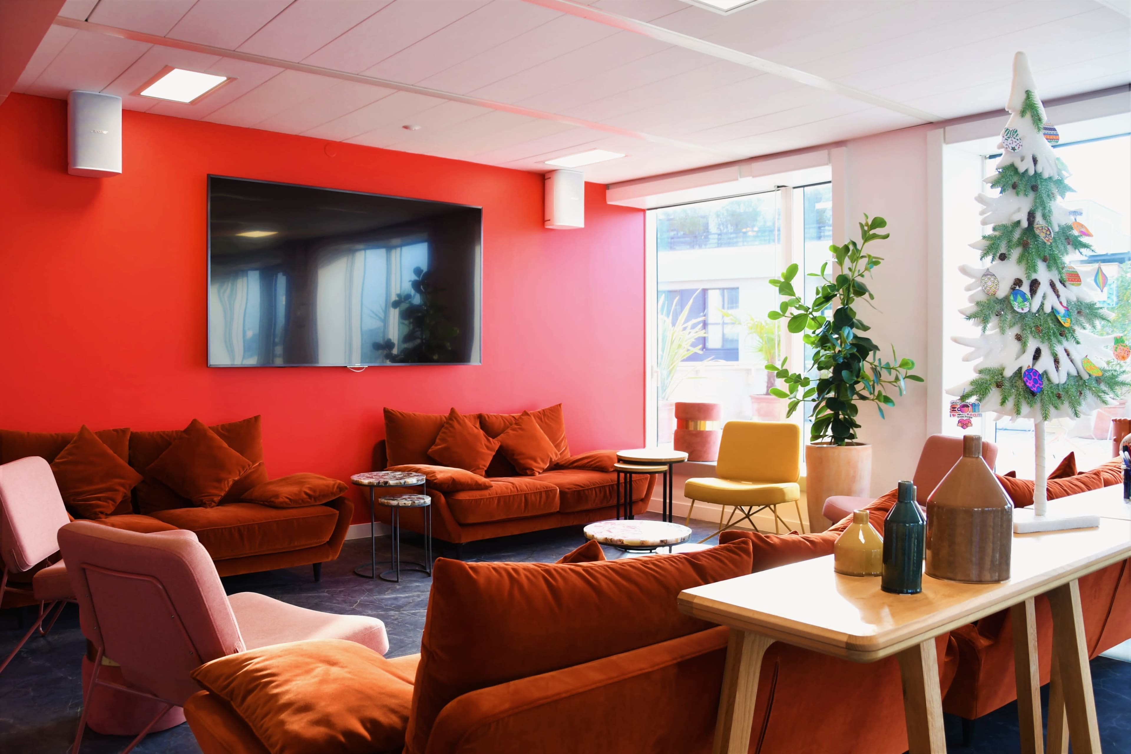 A modern lounge area with orange sofas, a large television mounted on a red wall, and a Christmas tree in one corner.