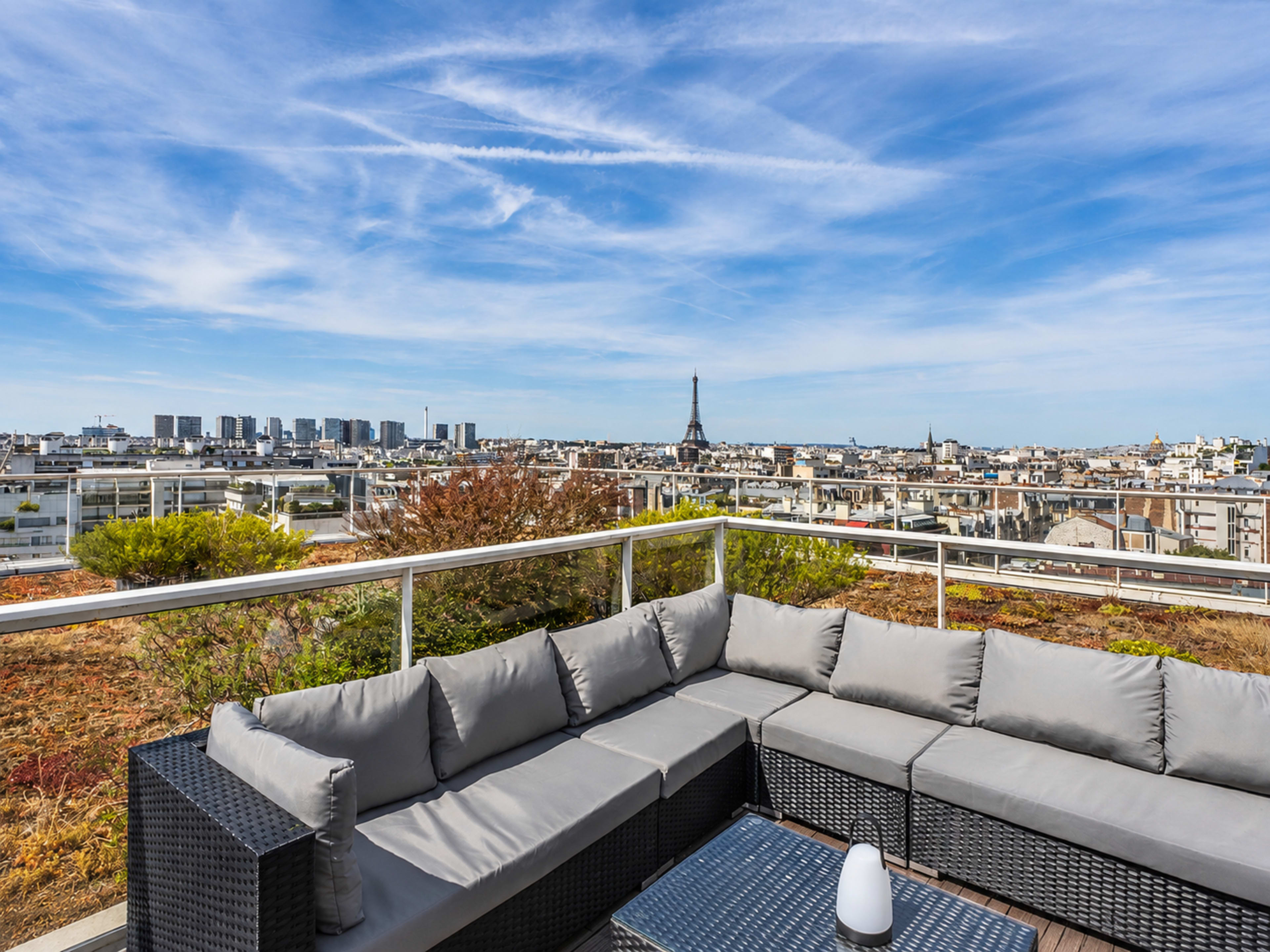 The image shows a rooftop terrace with a sectional sofa and coffee table overlooking a cityscape that includes the Eiffel Tower in the distance under a blue sky.