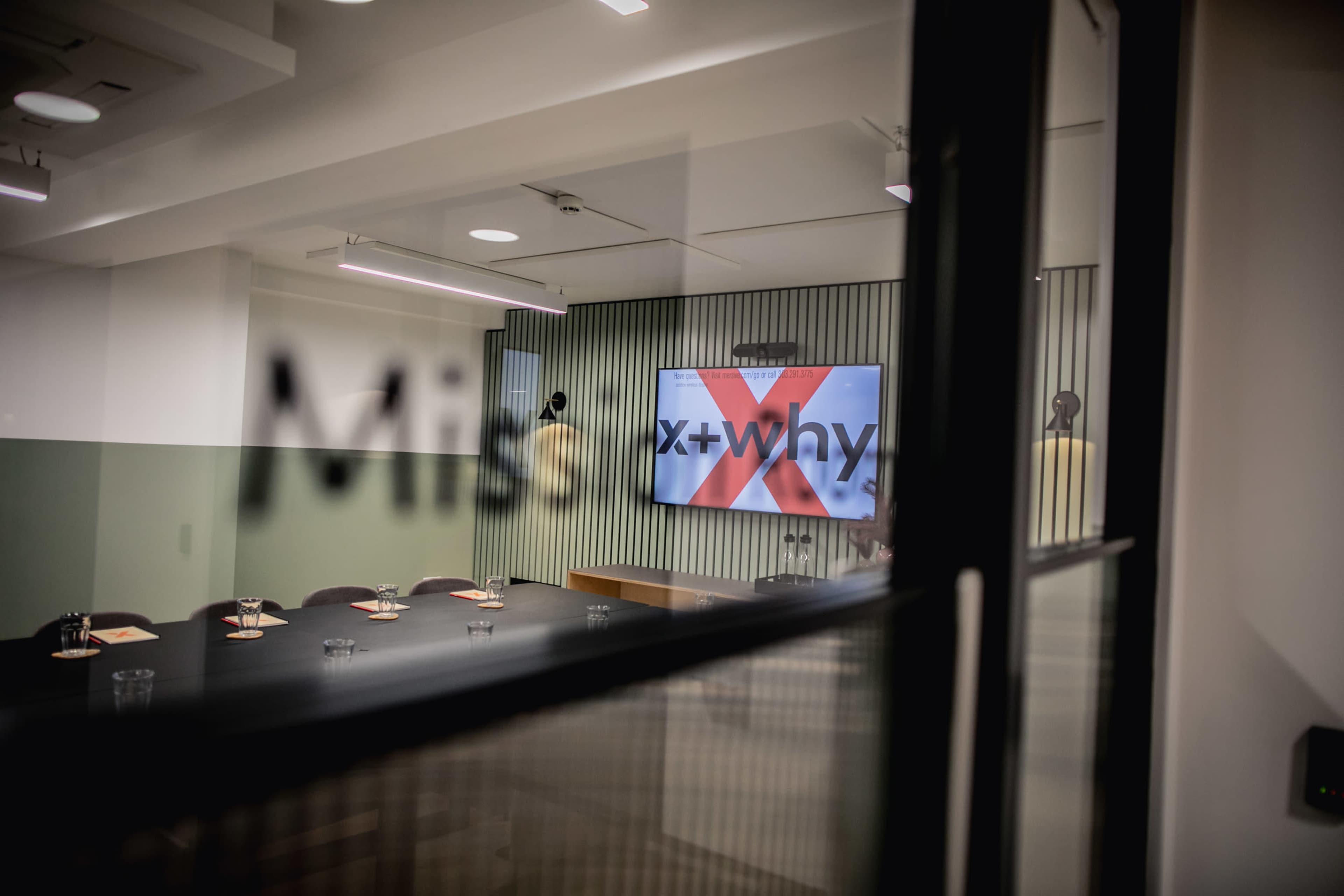 A conference room features a large screen displaying "x + why" and a table set with place cards.