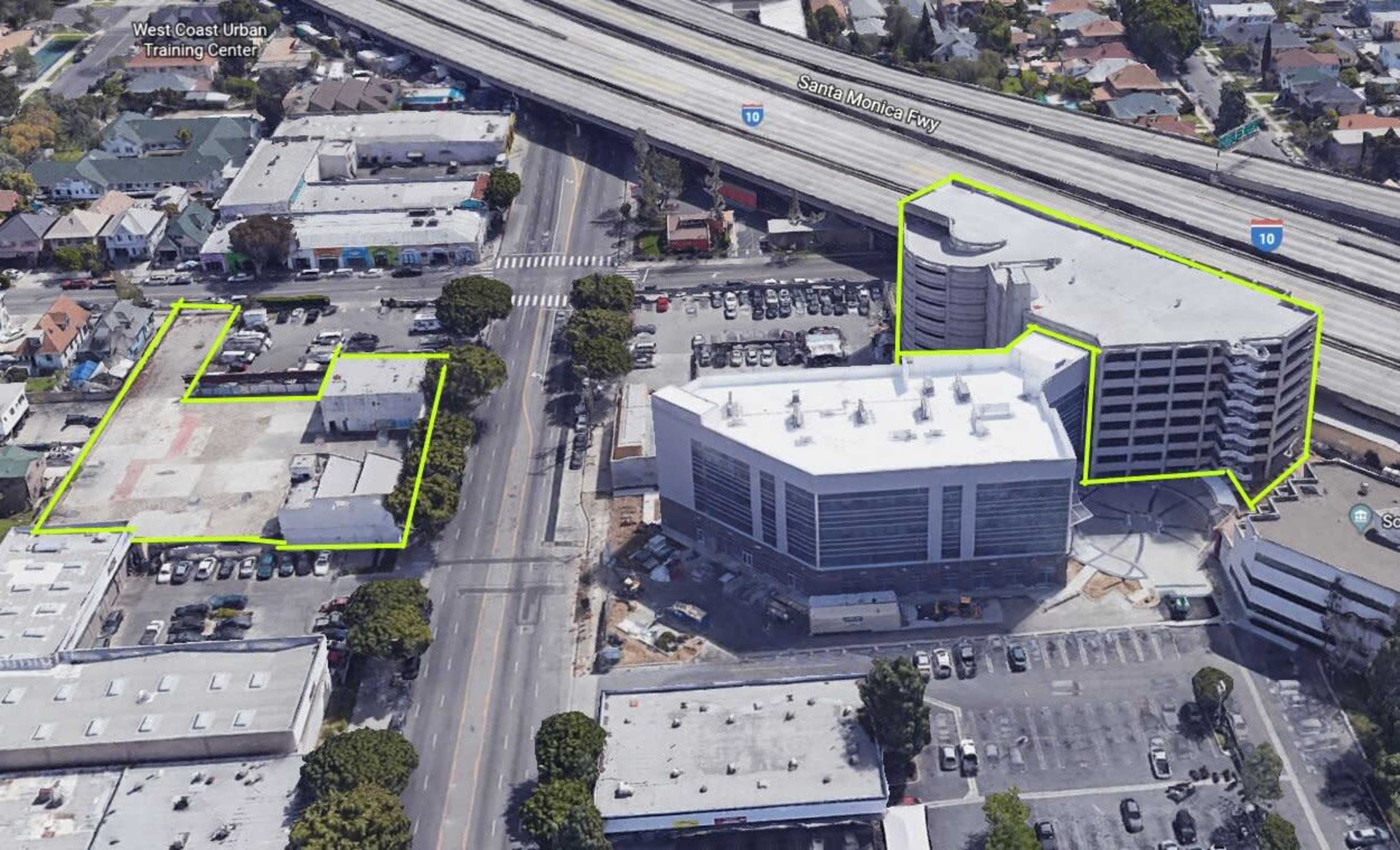 The image shows aerial view of a construction site featuring multiple commercial buildings and parking areas alongside a freeway.