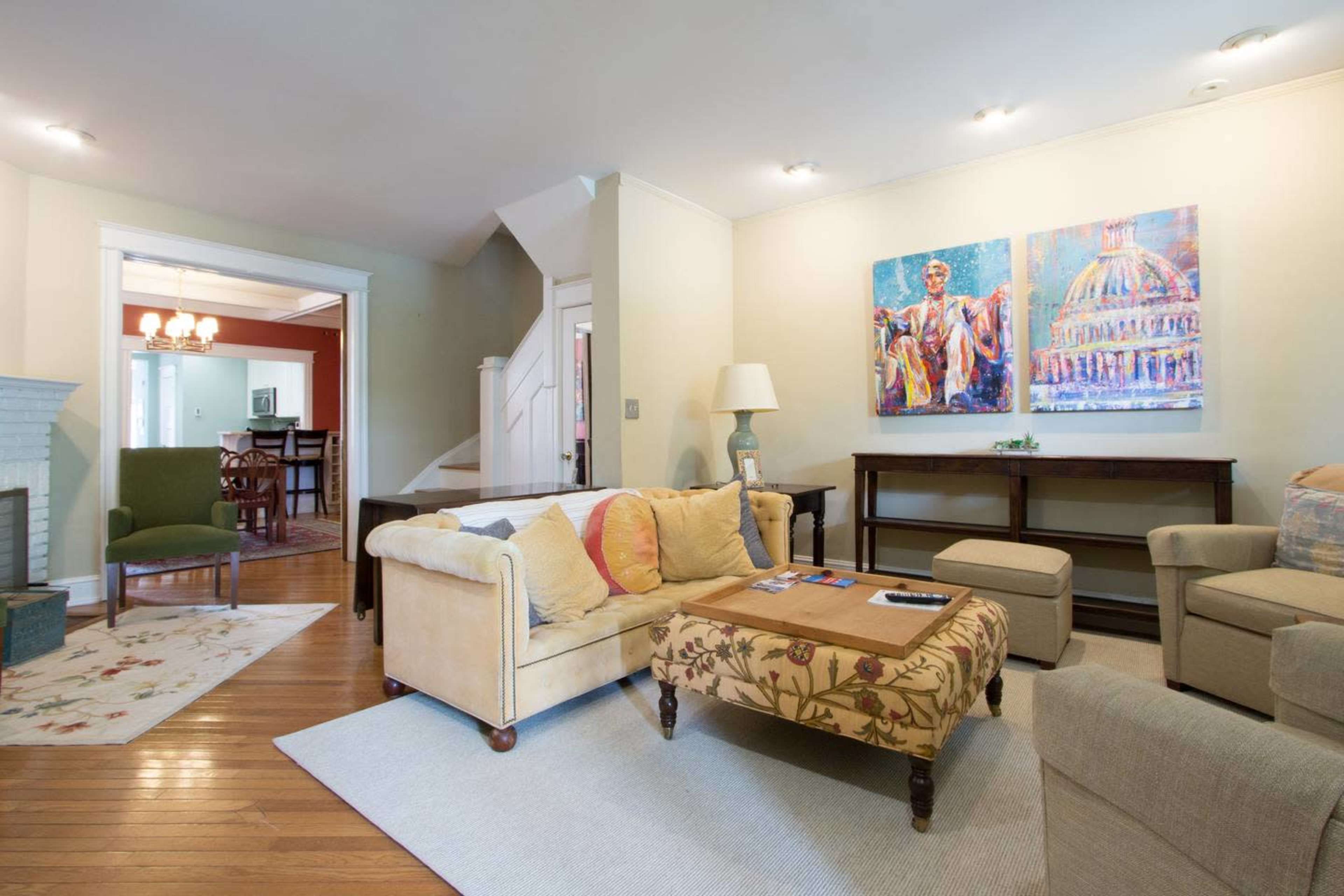 The living room features a light-colored sofa, a patterned ottoman, and artwork on the wall, with a view leading to a dining area in the background.