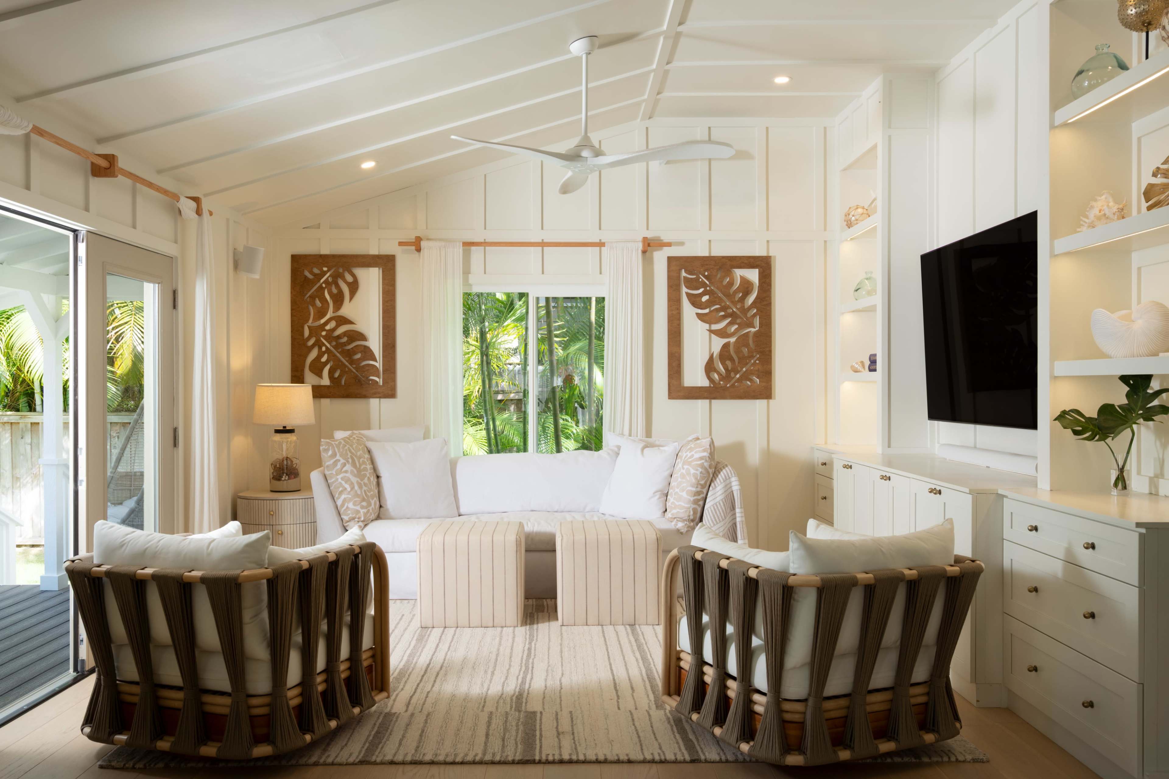 The image shows a bright, open living room featuring a white sofa, two patterned armchairs, and decorative wall art, with large windows providing a view of greenery outside.