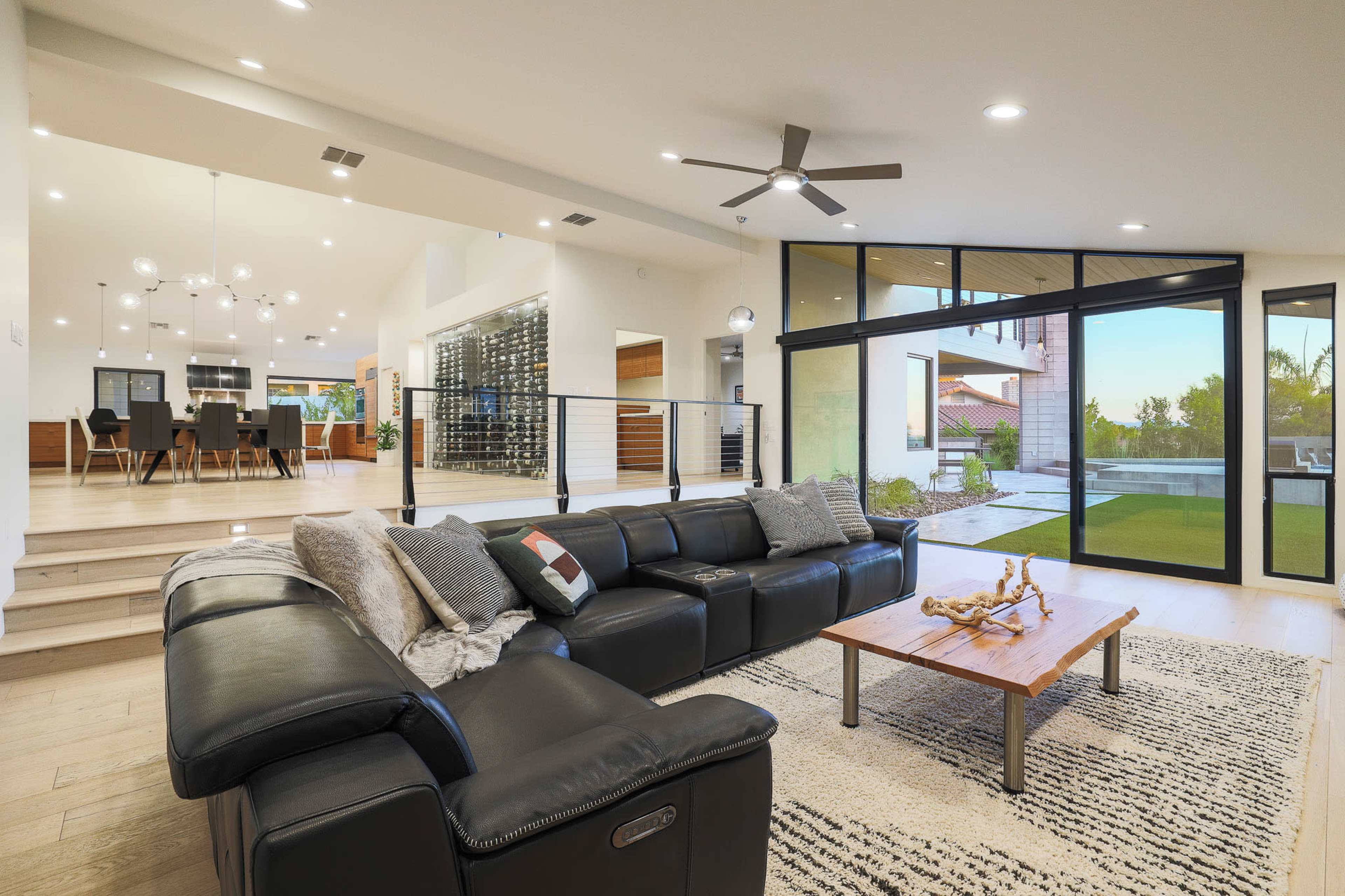 A modern living room with a black sectional sofa, a wooden coffee table, and large windows overlooking a patio and yard.