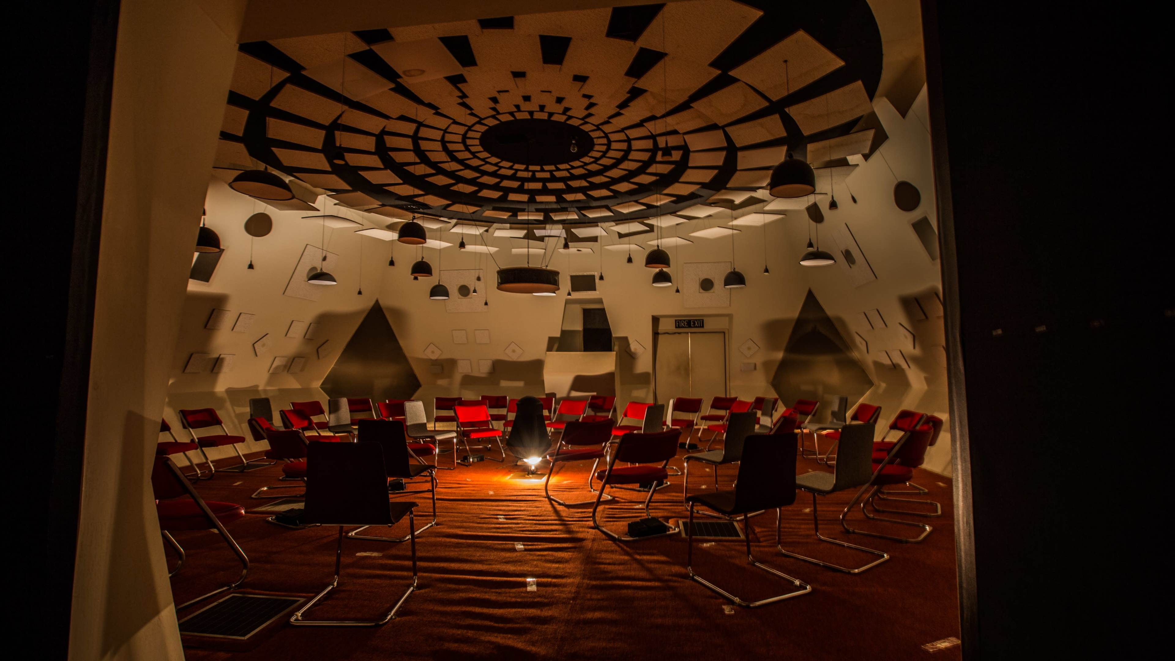 A circular room with a concentric ceiling pattern features red chairs arranged in a semicircle around a central light source.