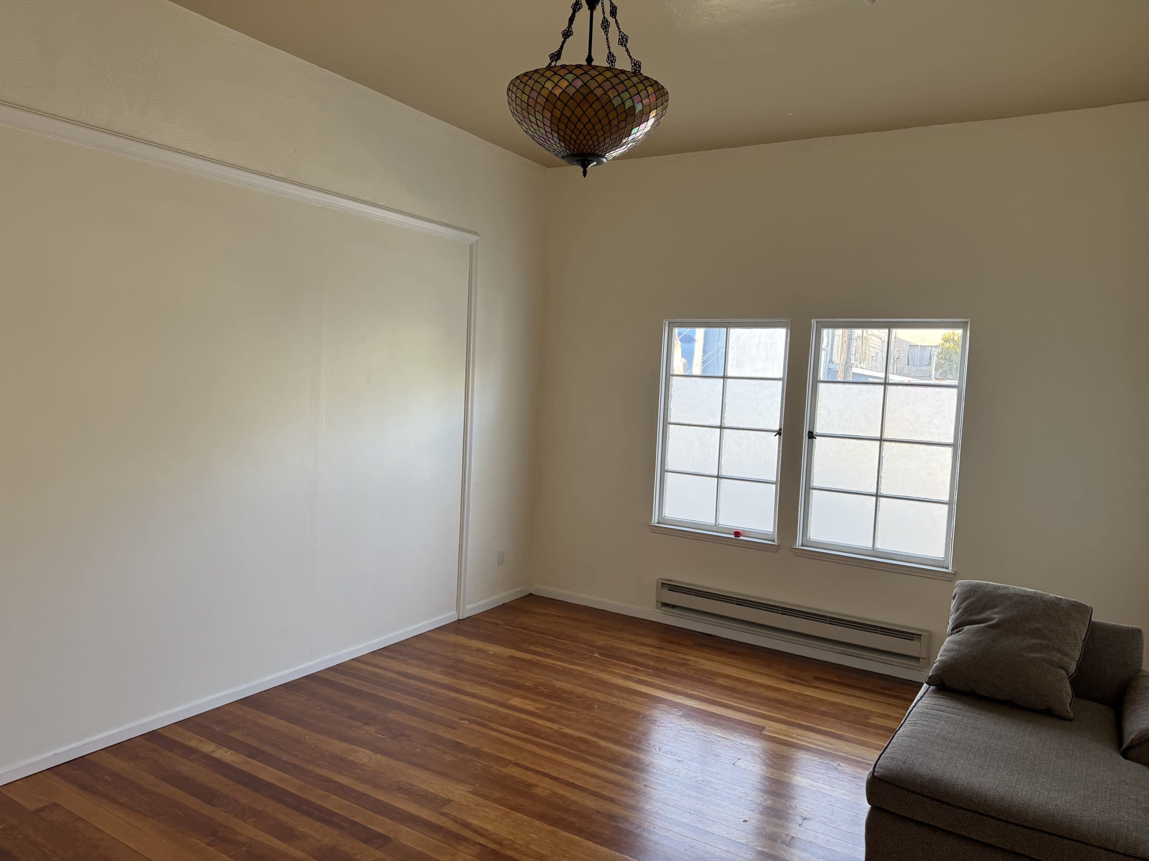 The image shows a bright, empty room with hardwood floors, a decorative light fixture, and two windows.