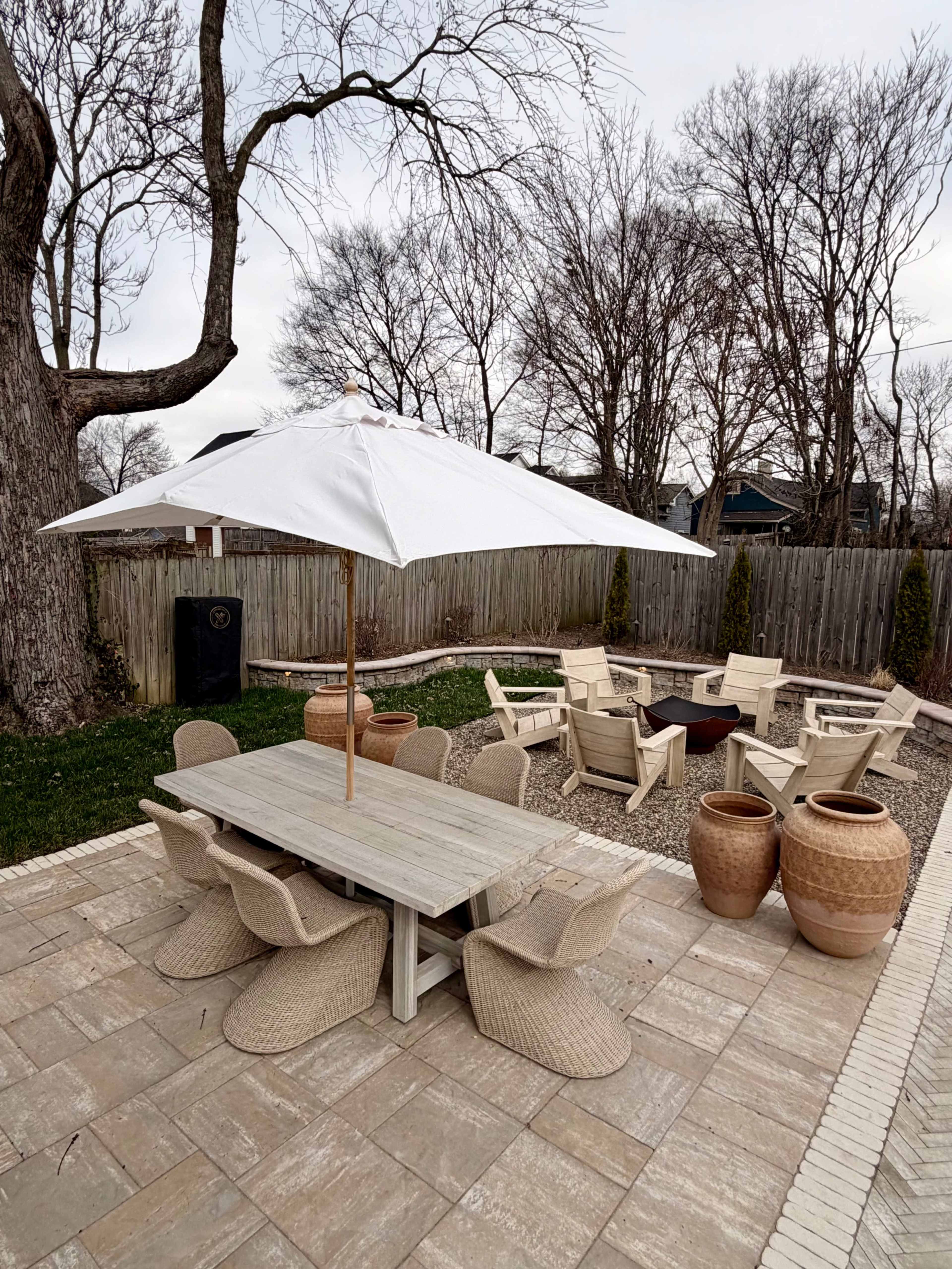 The image shows a backyard patio featuring a large table with an umbrella, surrounded by several chairs and two large planters.