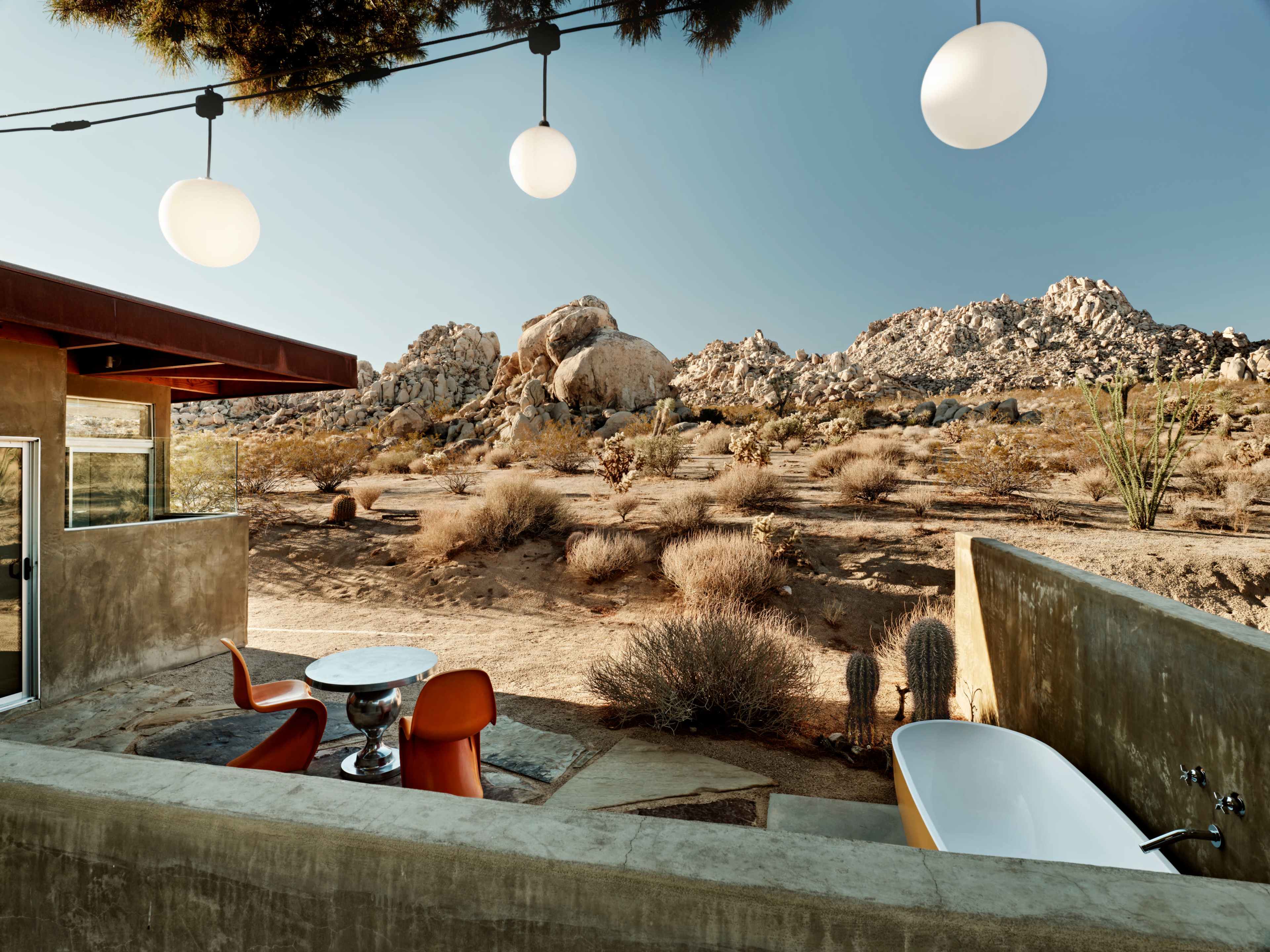 A modern outdoor space features a circular table with orange chairs, a freestanding bathtub, and boulders in the background under clear blue skies.