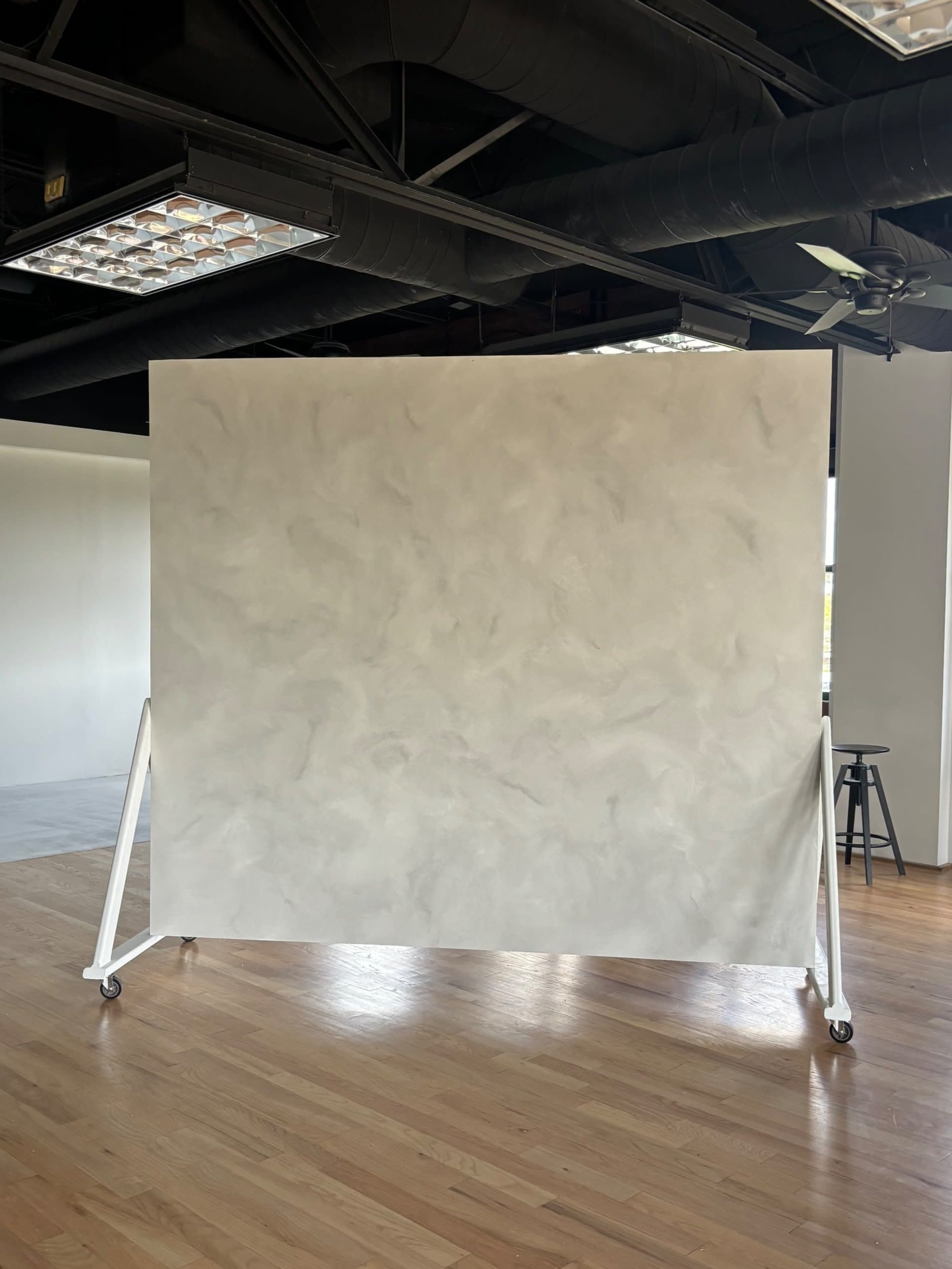 A large, smooth gray backdrop is mounted on a white, wheeled frame in a spacious, well-lit interior with wooden flooring.