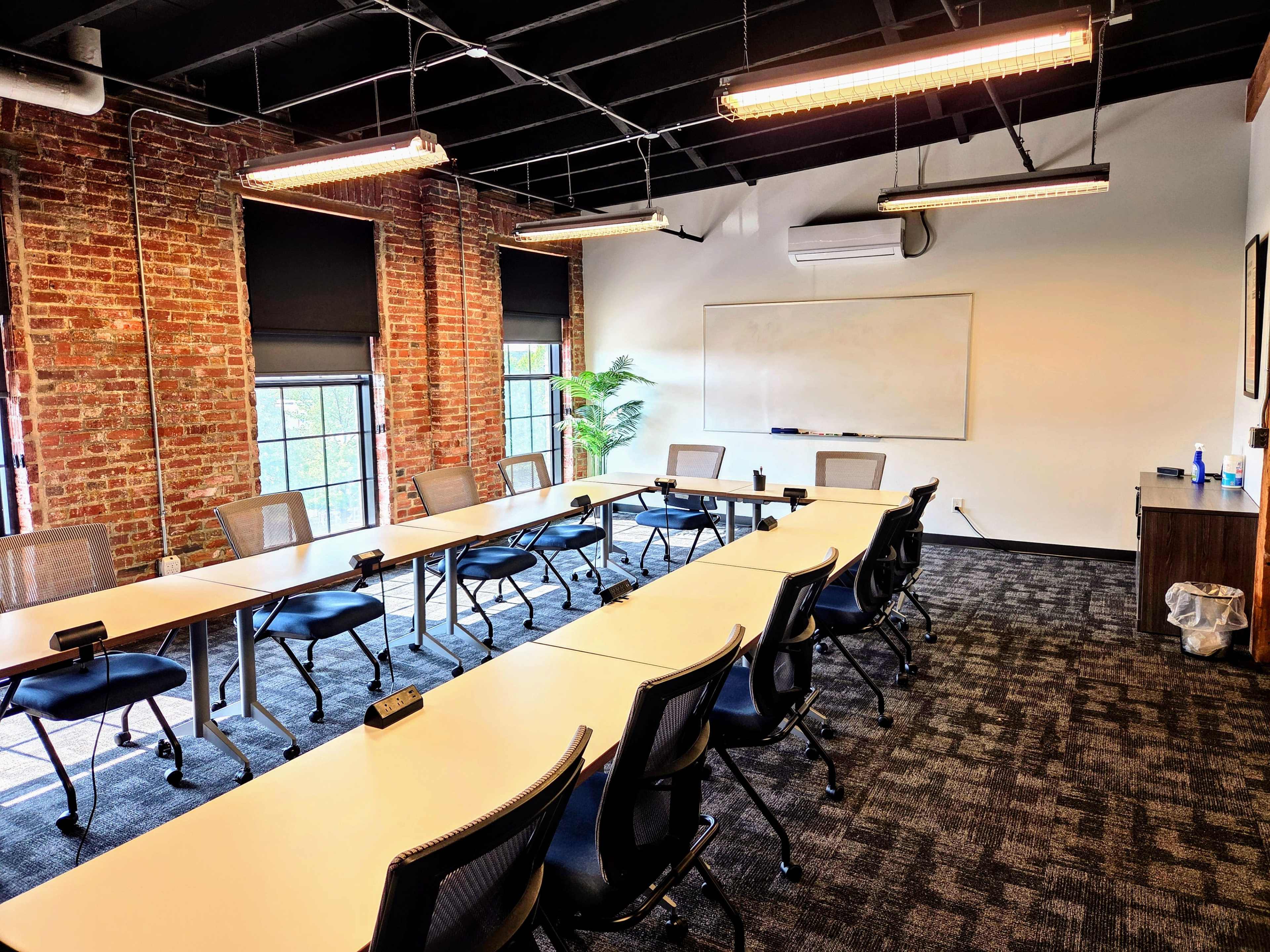 The image shows a conference room with a long rectangular table, several chairs arranged around it, exposed brick walls, and large windows letting in natural light.