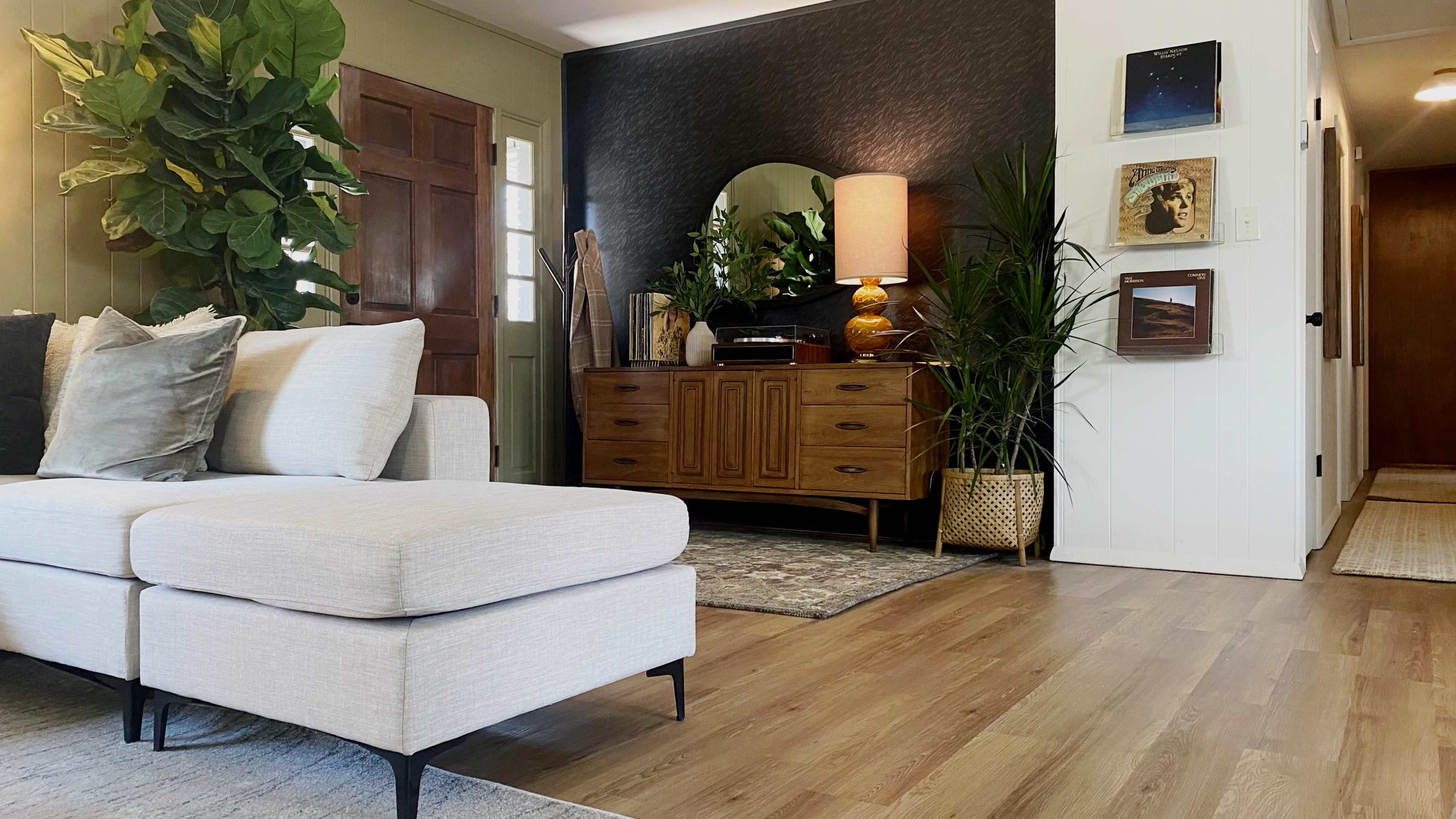 The image shows a living room with a light-colored sectional sofa, a wooden sideboard, and various indoor plants.