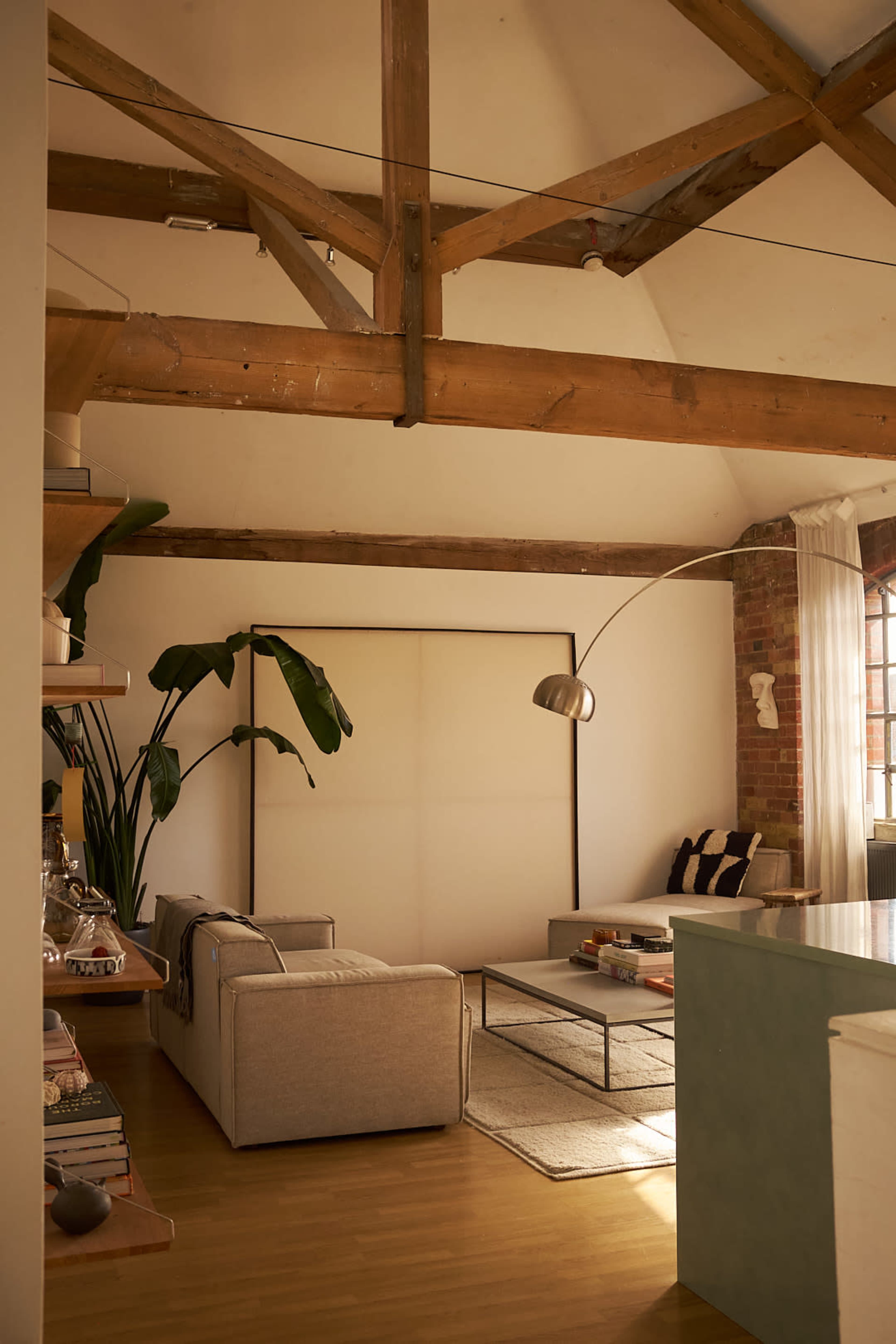 The image shows a spacious living room with wooden beams, a light-colored sofa, a glass coffee table, and a large plant in the corner.