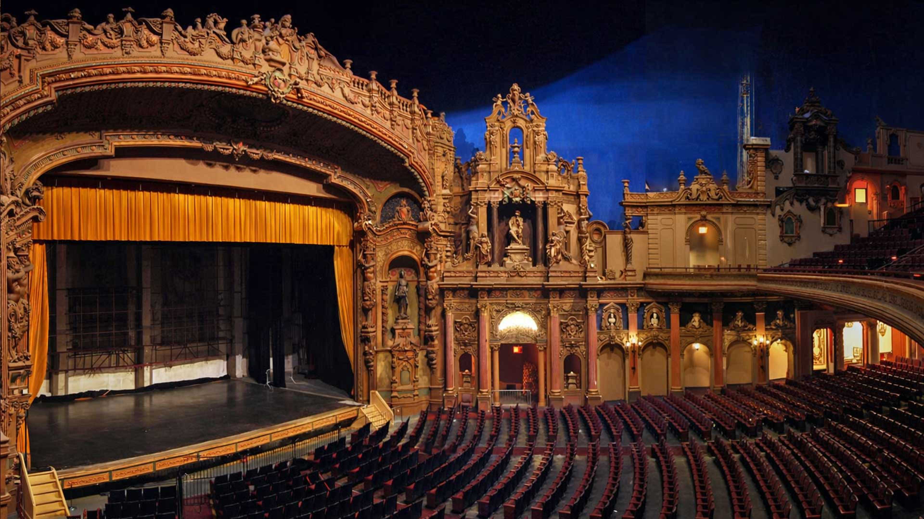 The image shows an ornate theater interior with a grand stage framed by intricate architectural details and a large, golden curtain.