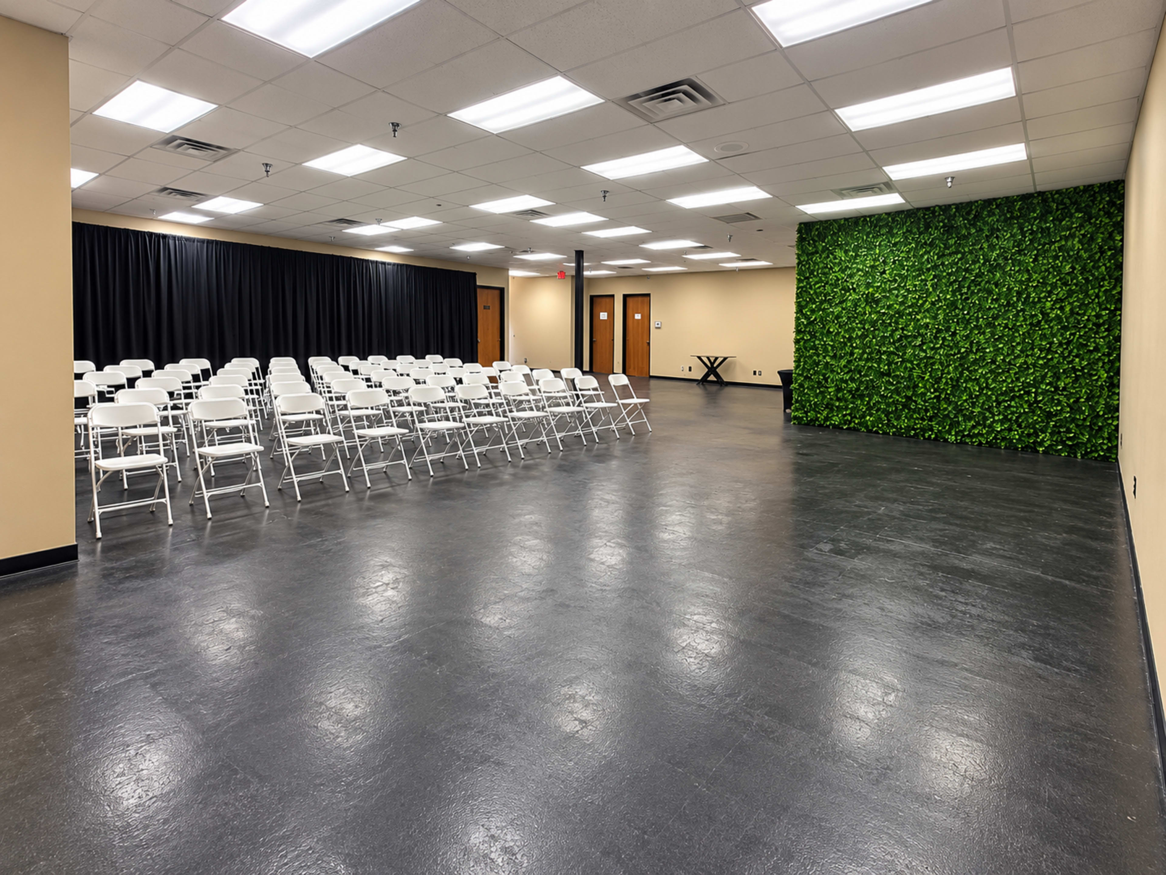The image shows an empty conference room with rows of white folding chairs facing a green accent wall and a black curtain backdrop.