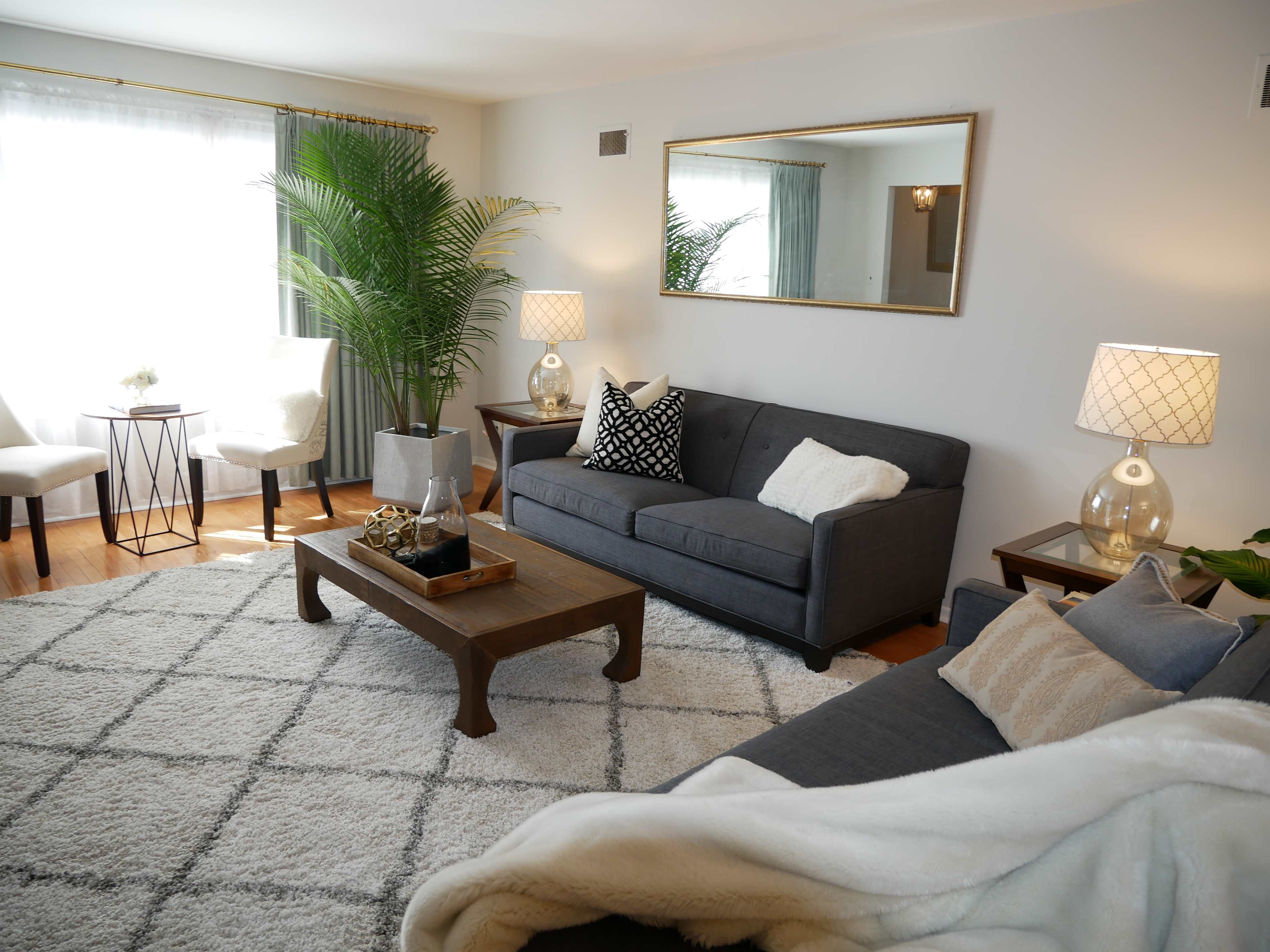 A living room features a gray sofa, a wooden coffee table, and a plush rug, with large windows allowing natural light and decorative plants enhancing the space.