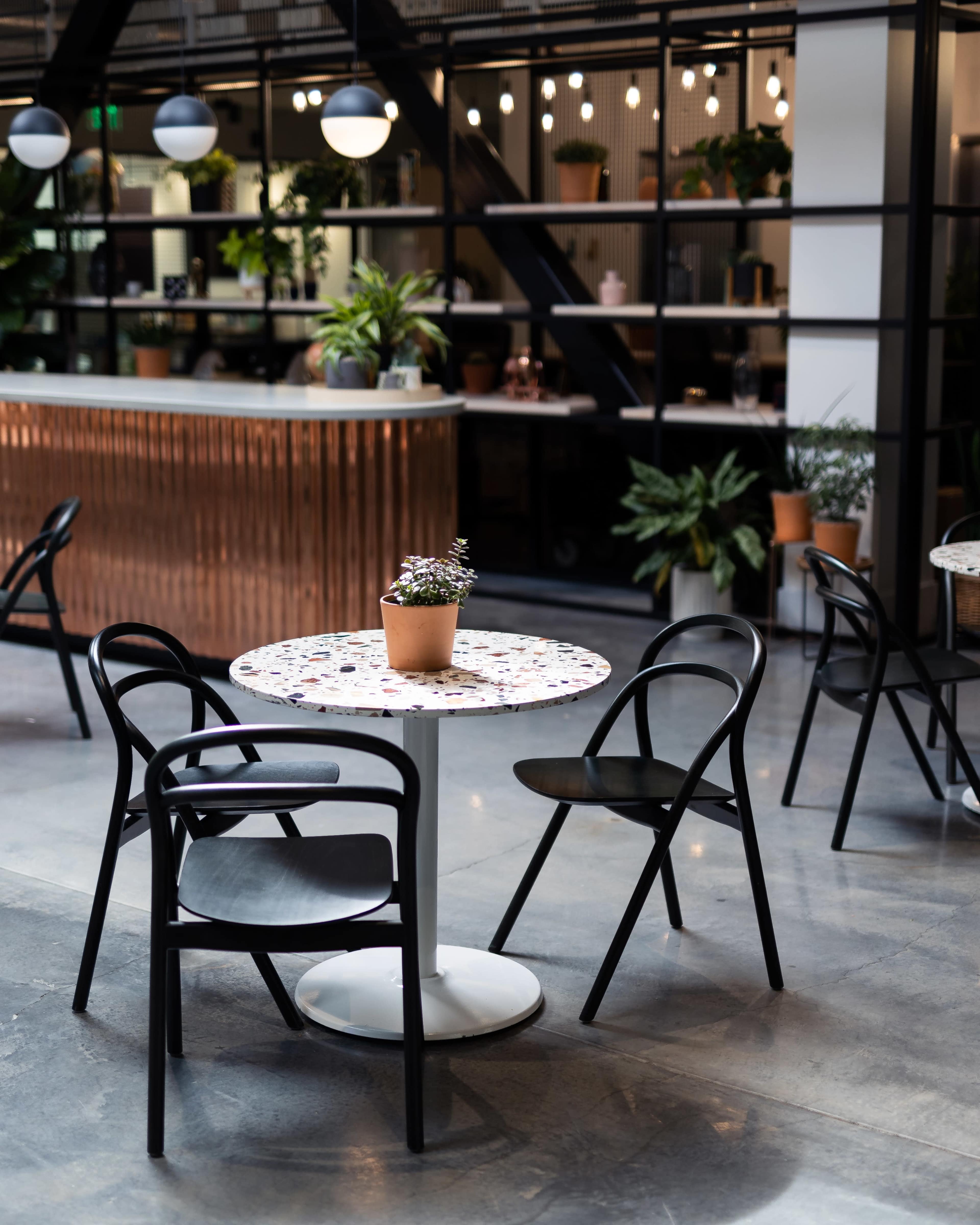 A small round table with a plant centerpiece is positioned among black chairs in a modern indoor café setting featuring greenery and a wooden bar.