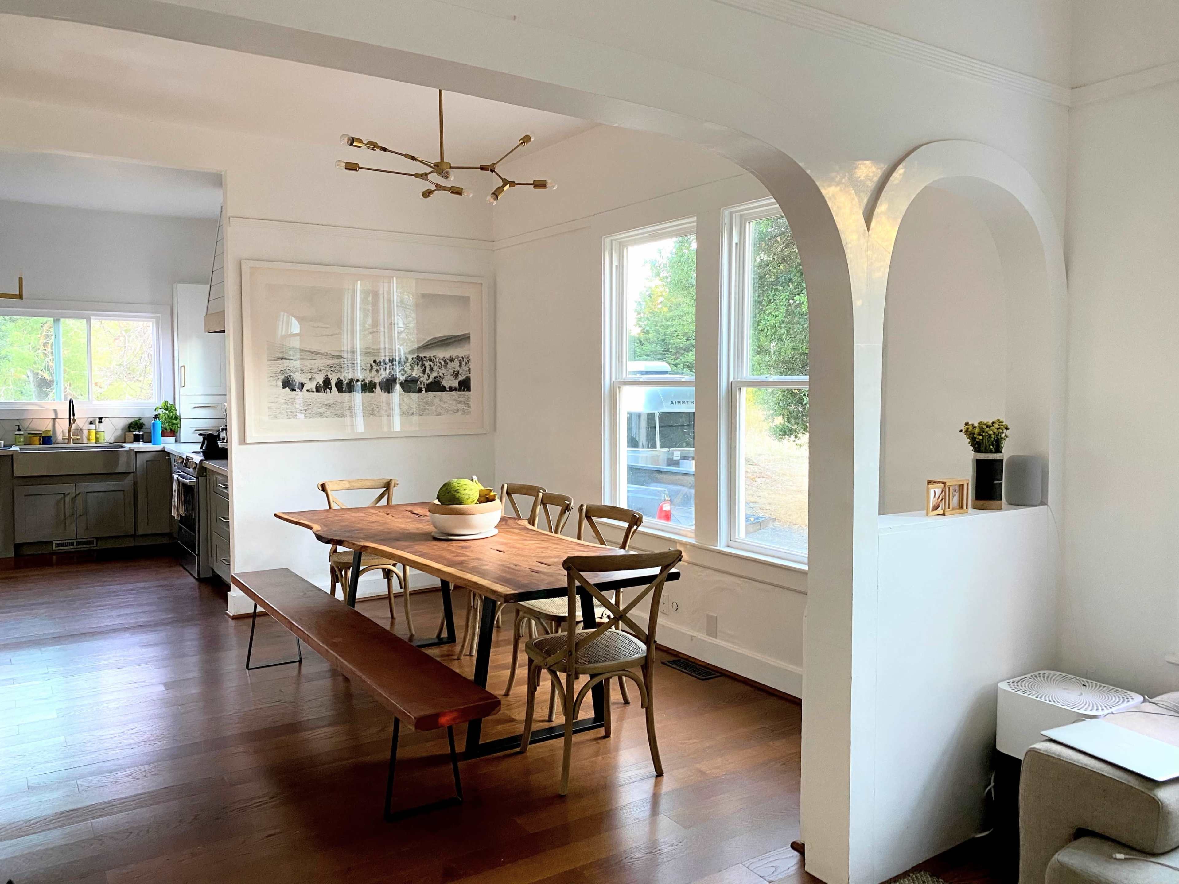 A dining area with a wooden table, several chairs, and large windows letting in natural light, featuring a minimalist design and a view of an outdoor area.