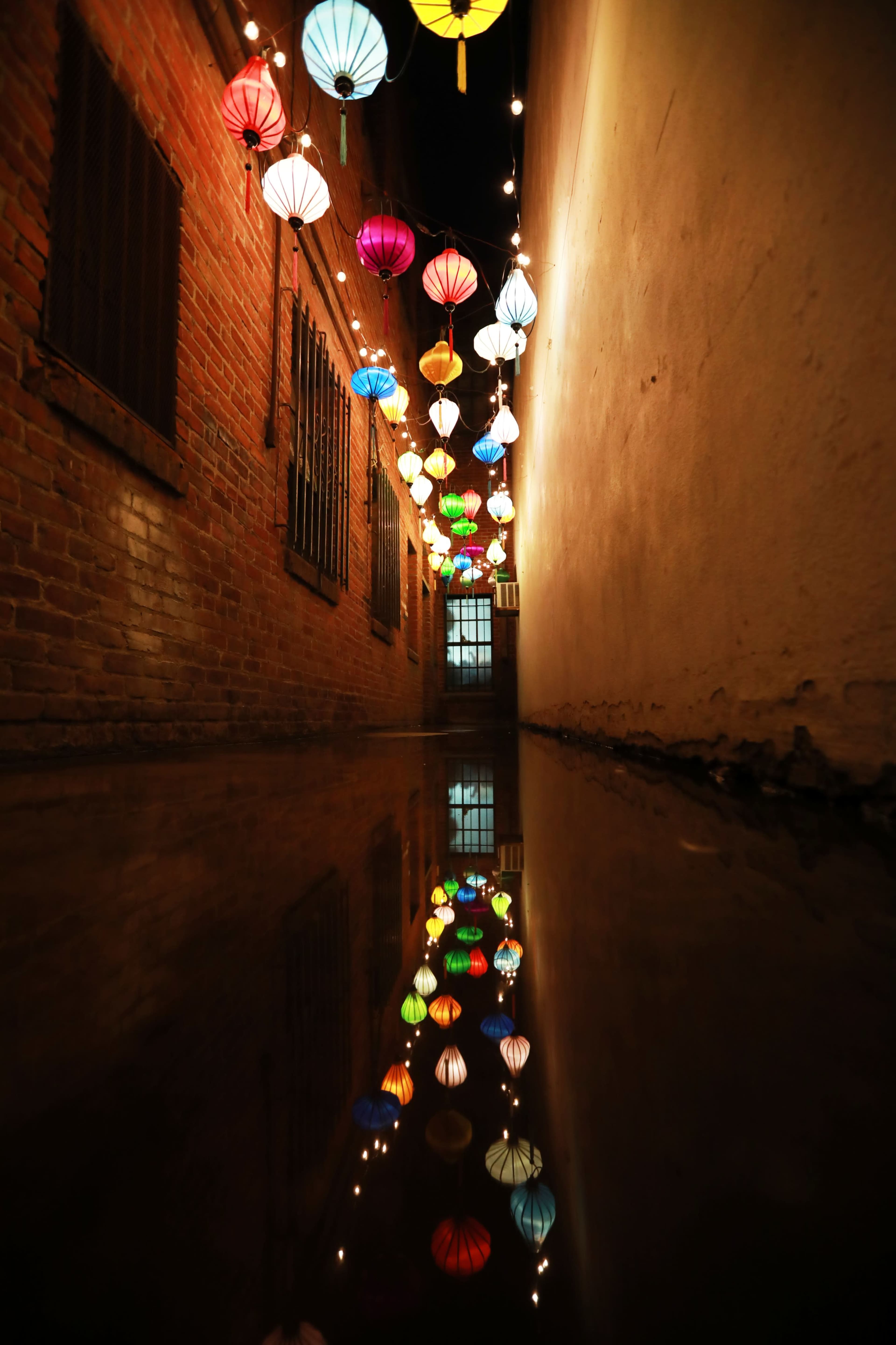 Colorful lanterns hang above a narrow alley, reflecting in a puddle on the ground.