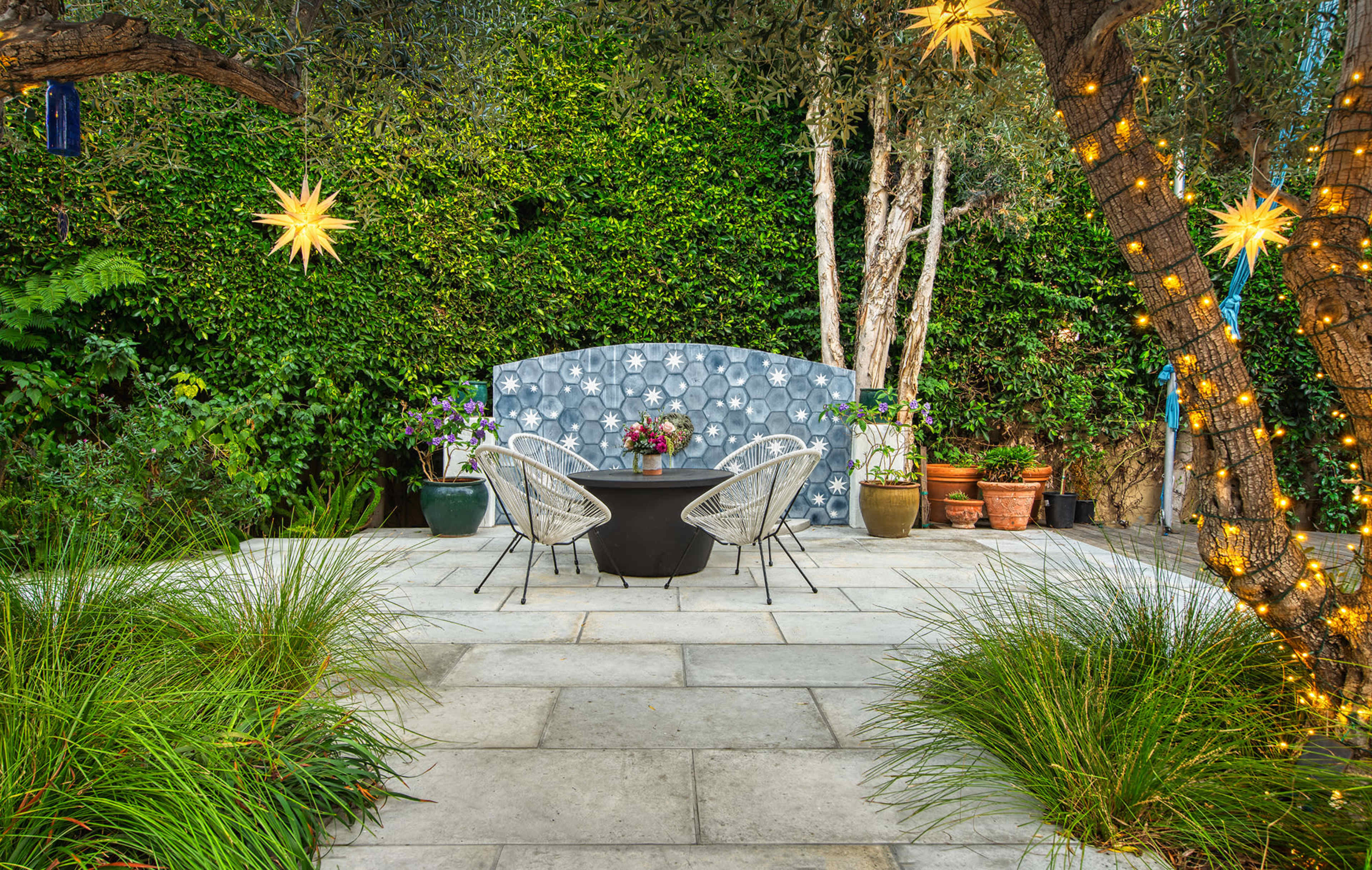 A stone patio features a round black table and two white chairs, surrounded by lush greenery and illuminated by star-shaped lights hanging from the trees.