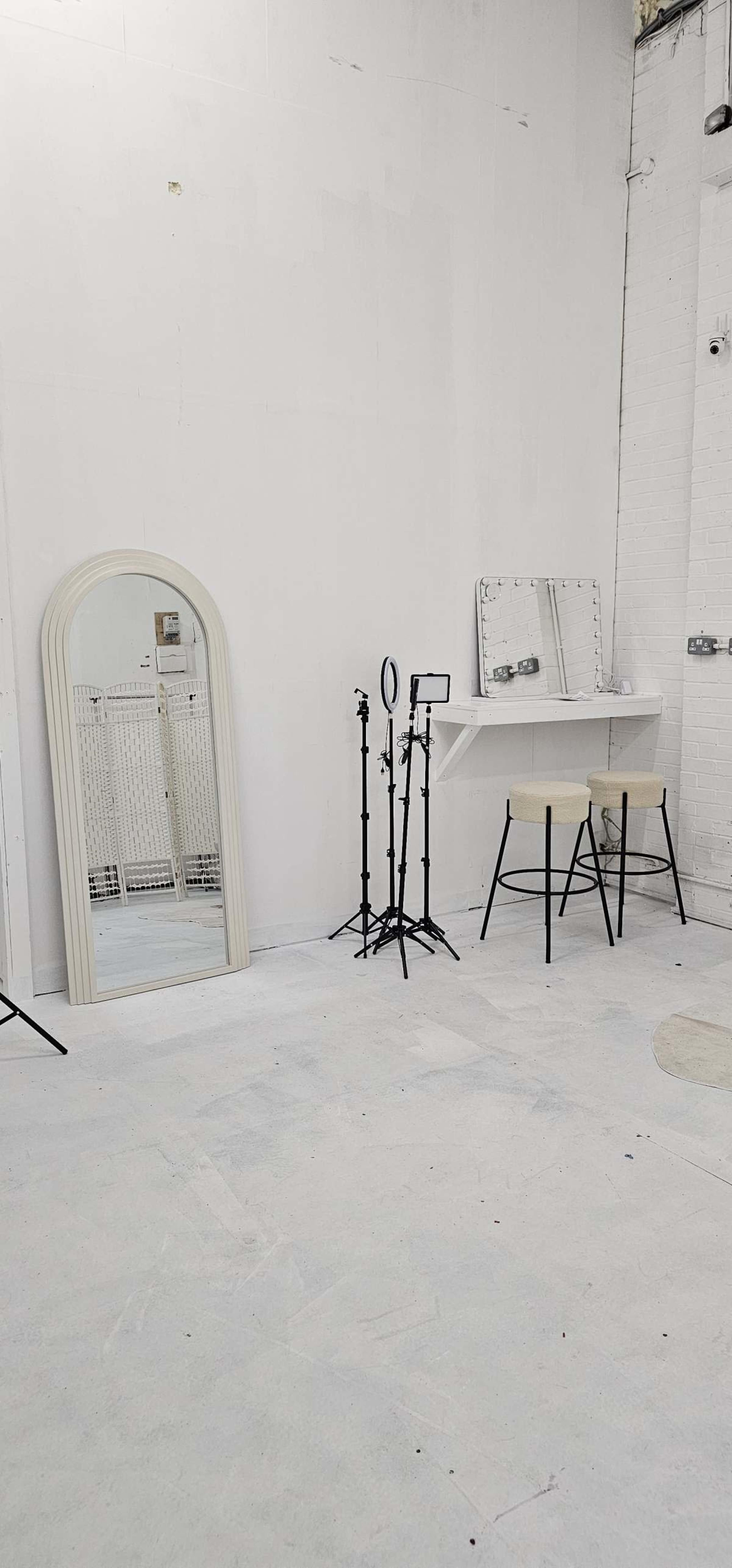 The image shows a minimalist indoor space with white walls, featuring a large mirror, a lighted mirror on a makeup table, and two tall stools.