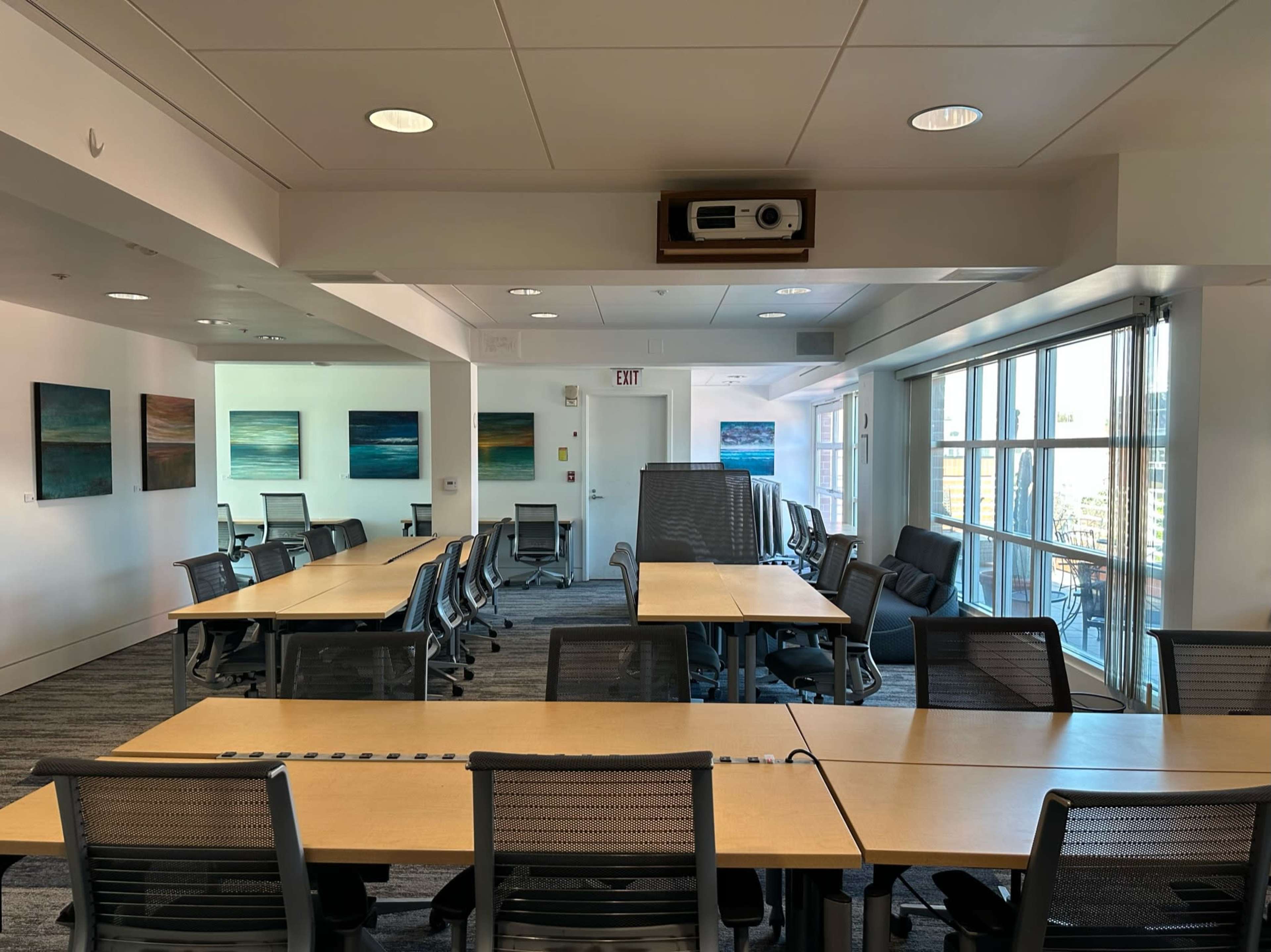 A spacious meeting room with rows of tables, chairs, and artwork on the walls, illuminated by natural light from large windows.