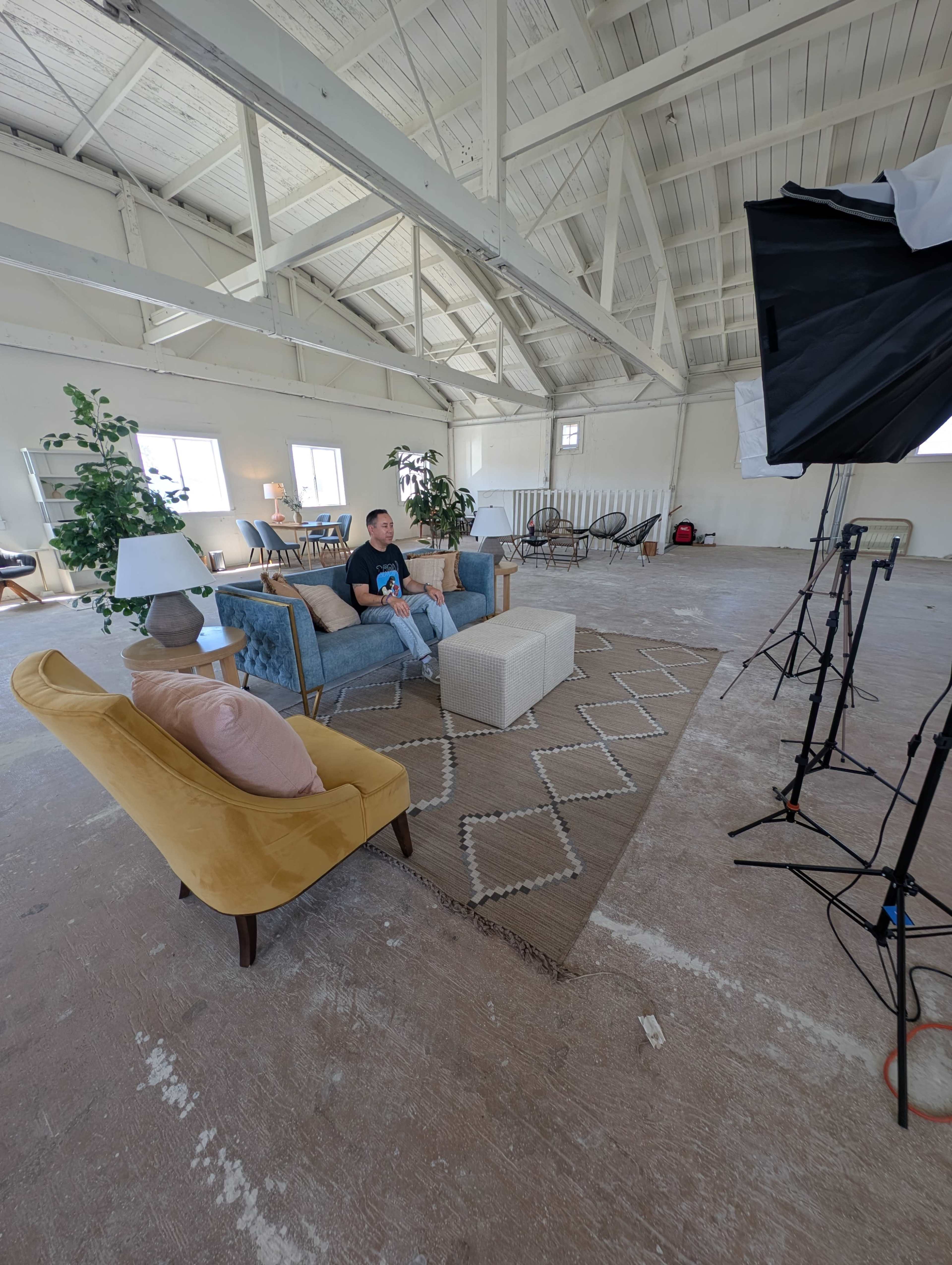 A man sits on a blue couch in a spacious room with large windows, surrounded by plants and photography equipment.