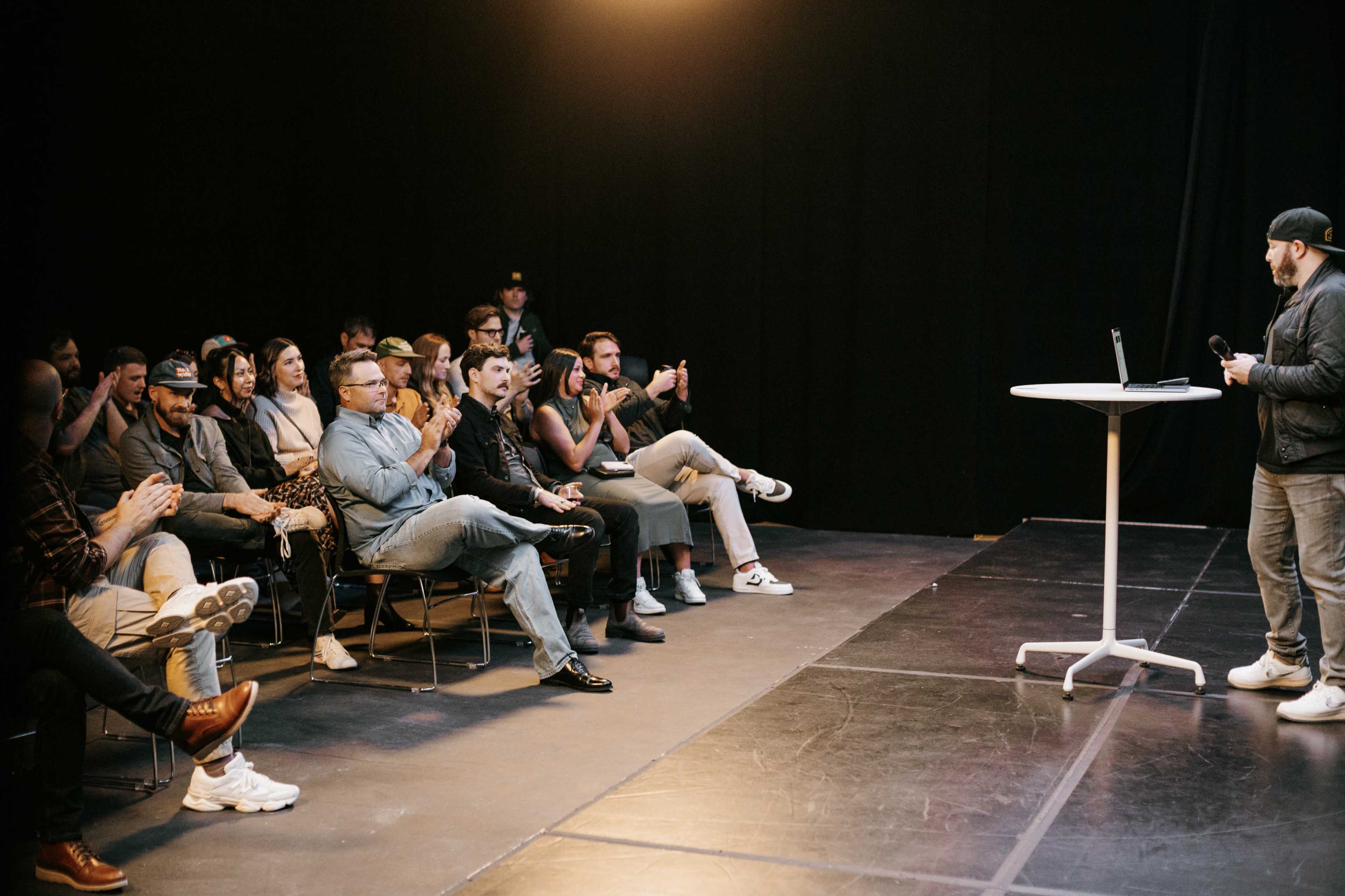 A speaker stands on a stage with a laptop while an audience claps and uses their phones.