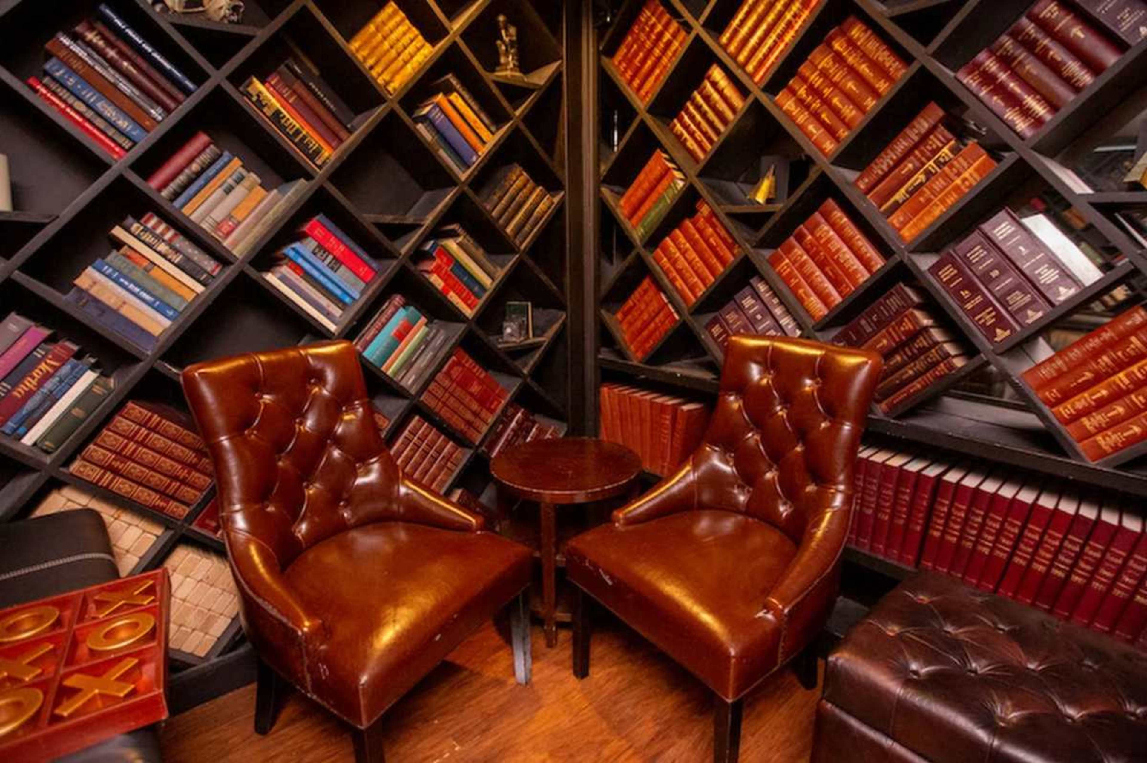 The image shows a cozy reading nook with two brown, tufted chairs flanking a small round table, set against a backdrop of neatly arranged bookshelves filled with colorful books.
