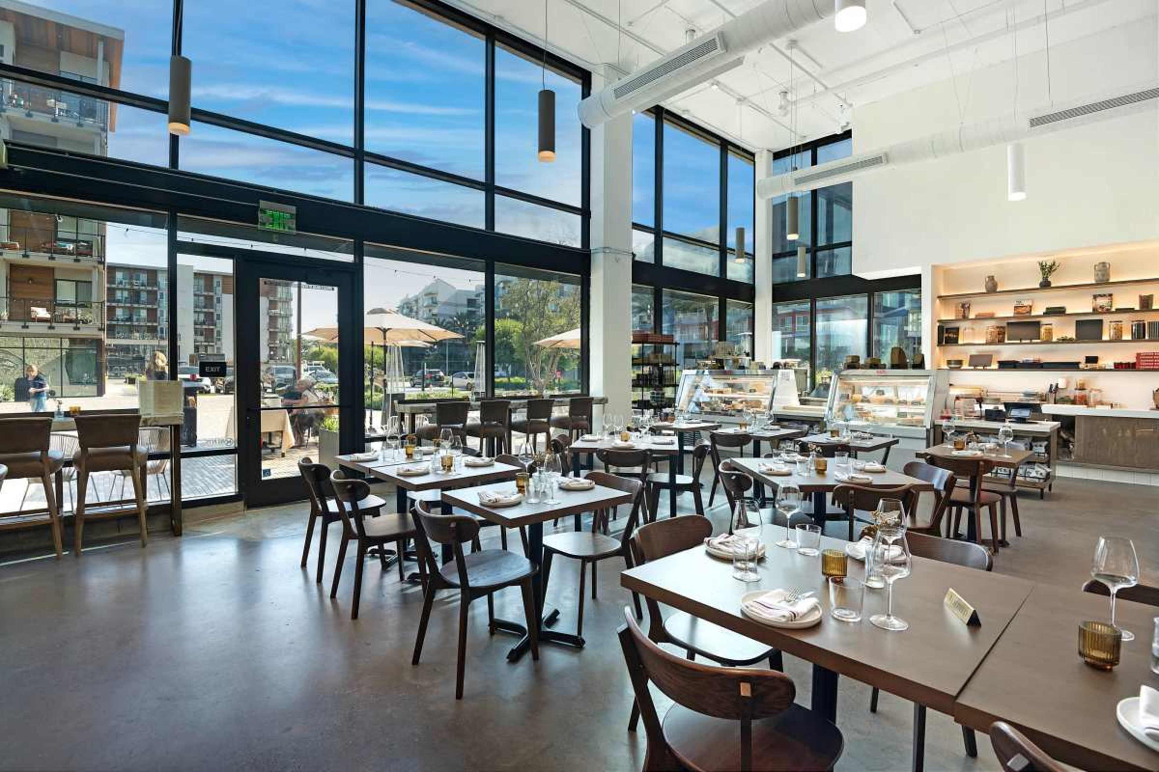 The image shows a modern, well-lit restaurant interior featuring tables set for dining, large windows with views outside, and a display of food items in a glass case.