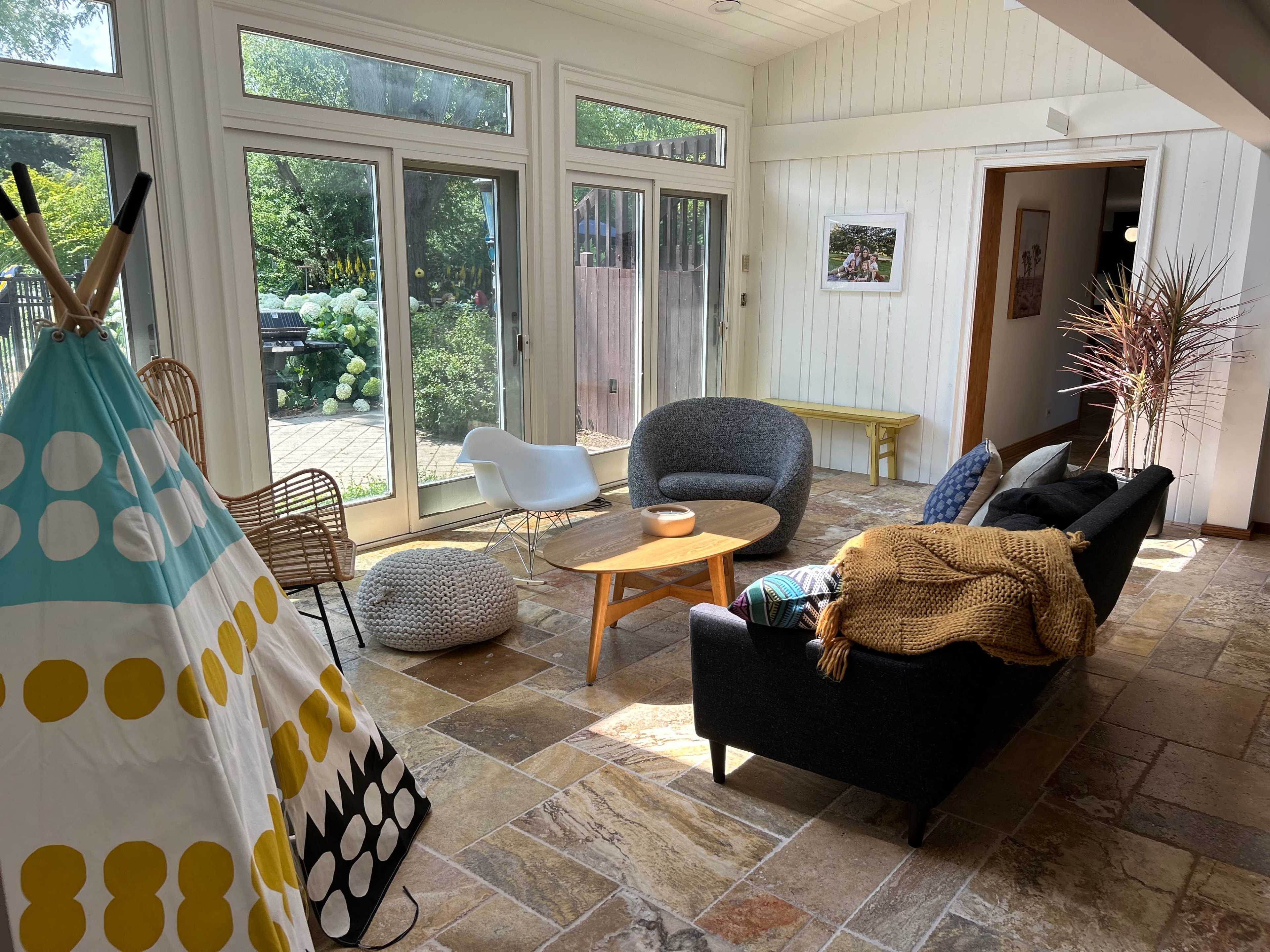 A cozy living room with a mix of seating options, including a sofa, armchairs, and a teepee, surrounded by large windows that let in natural light.