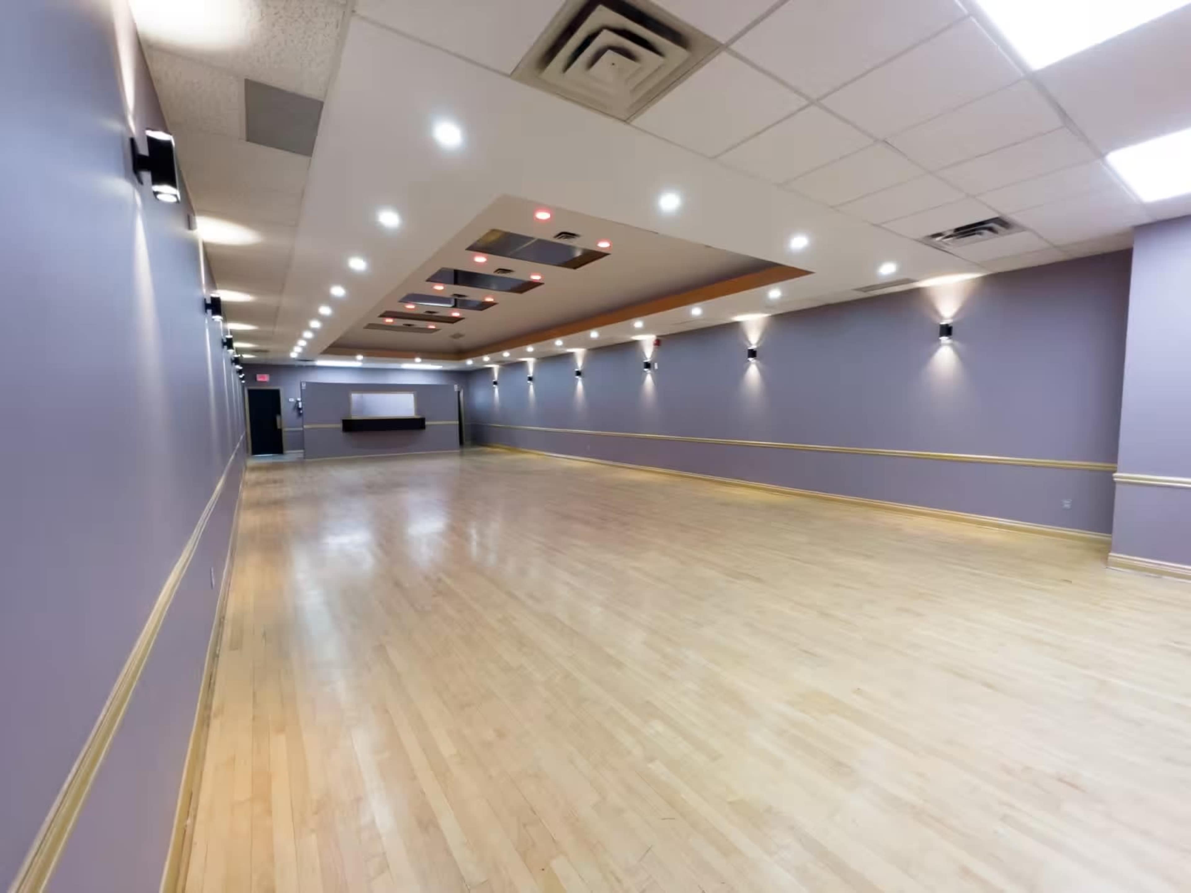A spacious dance studio with polished wooden flooring and purple walls, featuring recessed lighting and a front reception area.