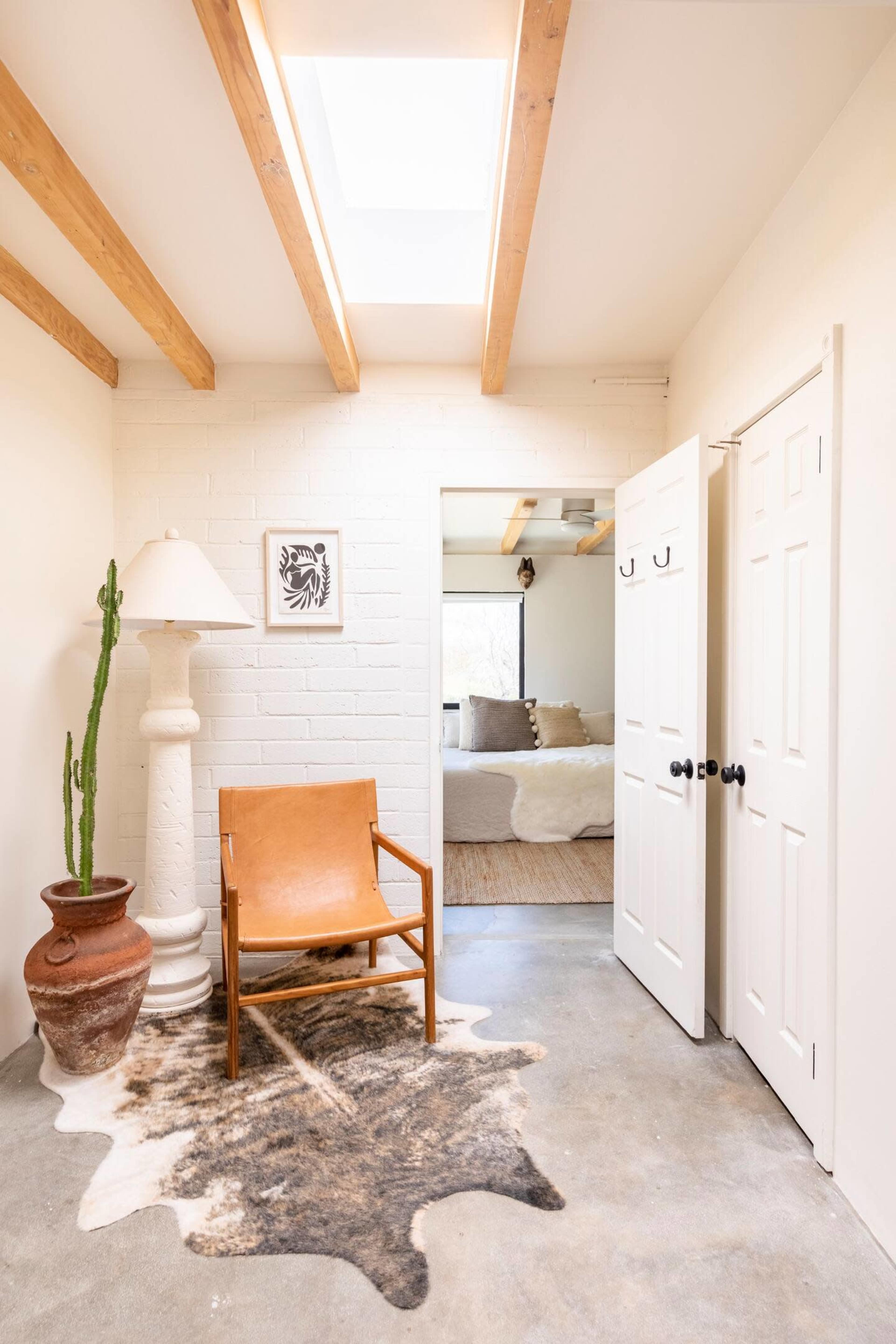 A bright interior hallway features a wooden-beamed ceiling, a chair, a lamp, a potted cactus, and a view into a softly decorated bedroom.