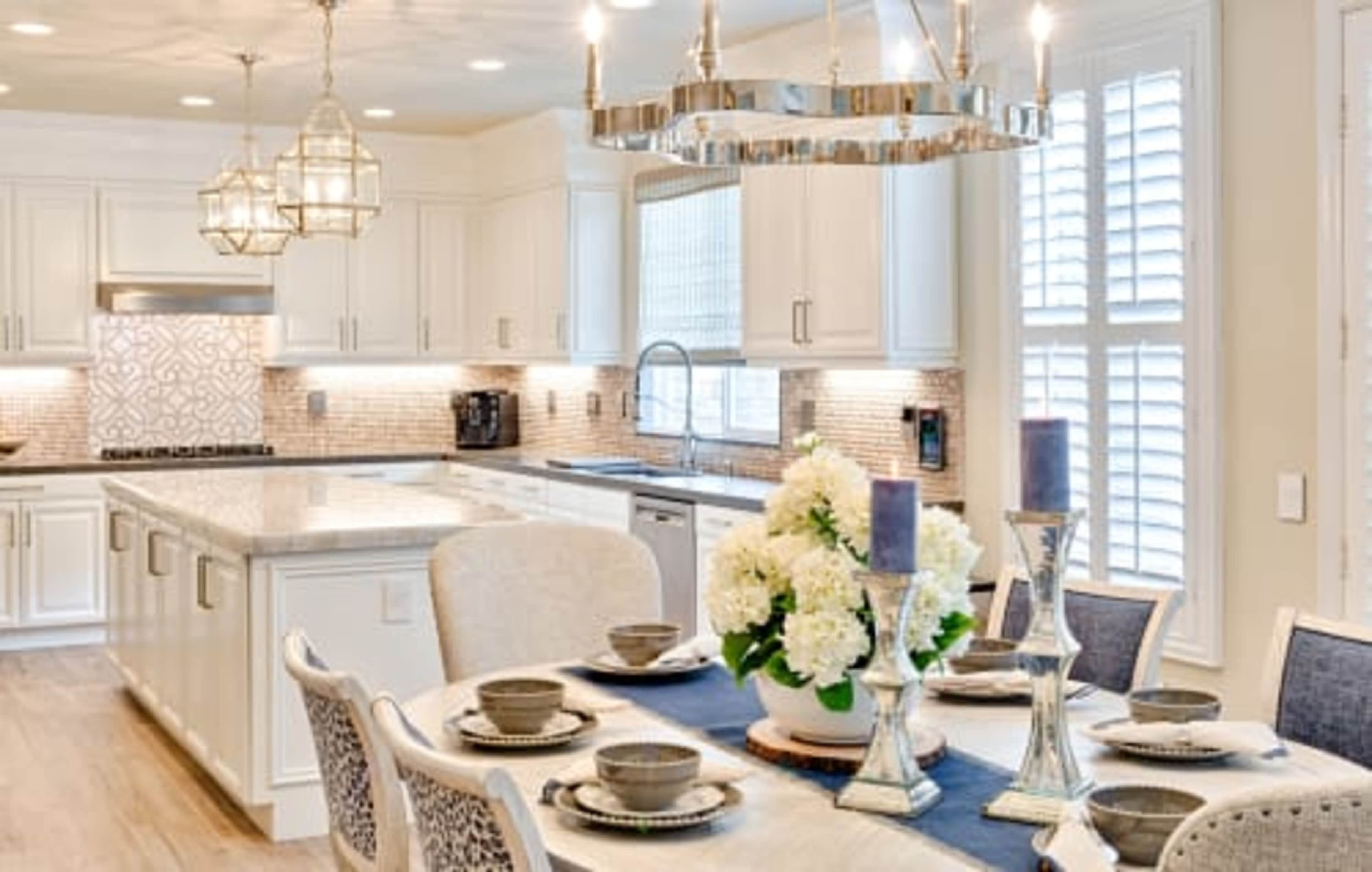 A modern kitchen and dining area features a table set for a meal, with a bouquet of white flowers and candles, surrounded by elegant decor and ample natural light.
