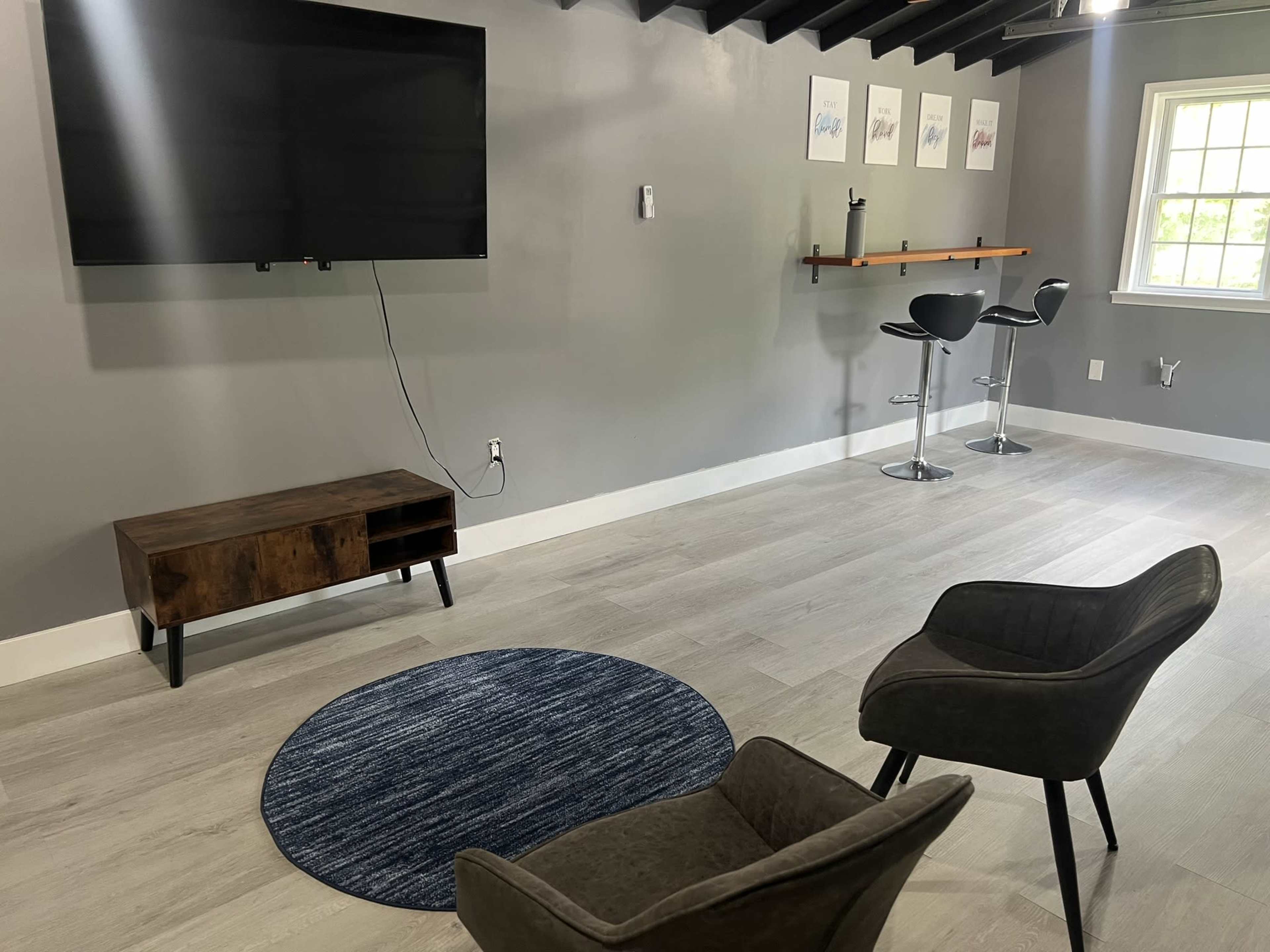 A room with a large flat-screen TV mounted on the wall, two gray chairs facing a circular blue rug, and a bar area with two black stools against a gray wall.