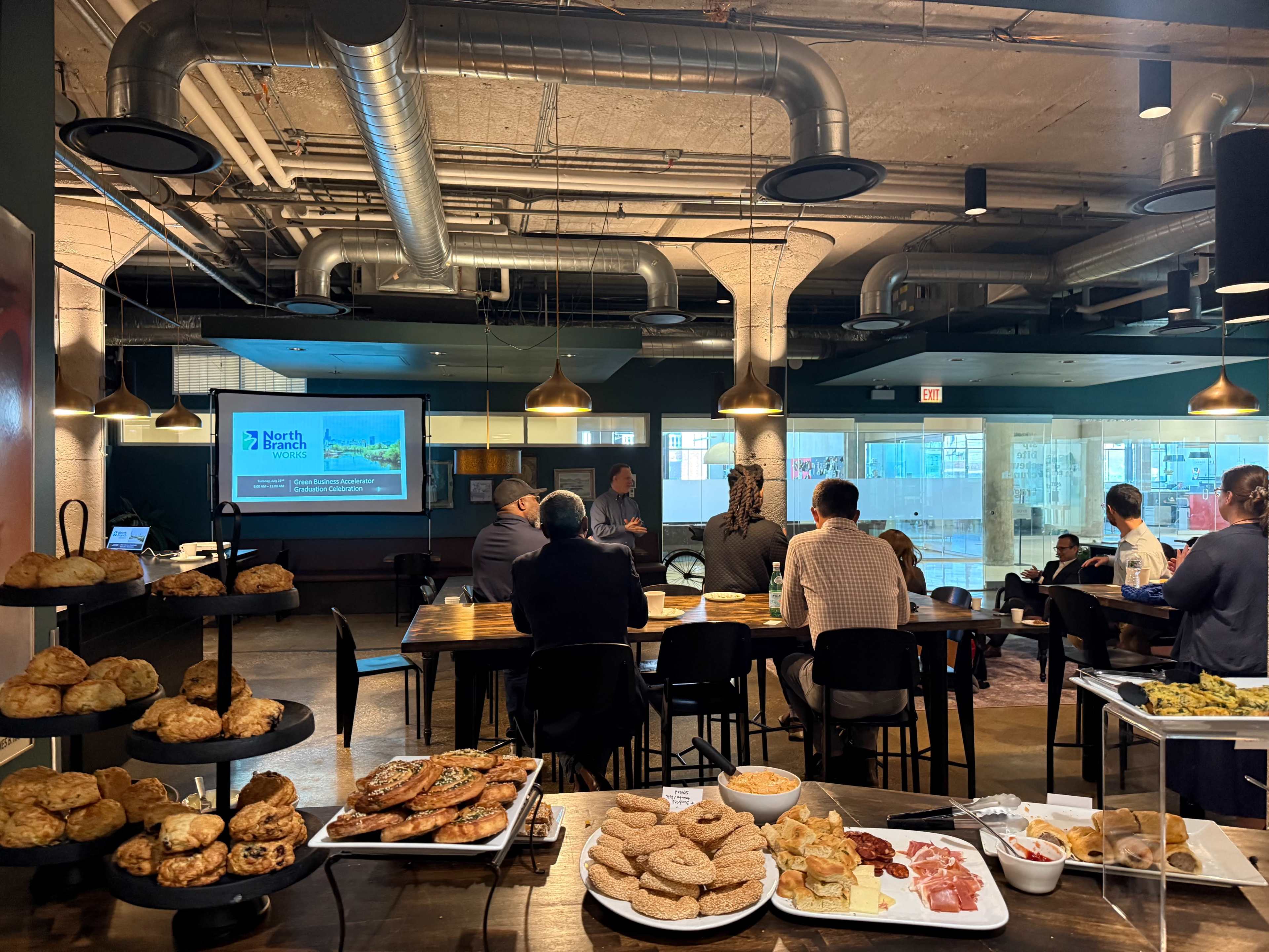 A group of people is seated in a modern meeting space with food laid out on a table, while a presentation is displayed on a screen.
