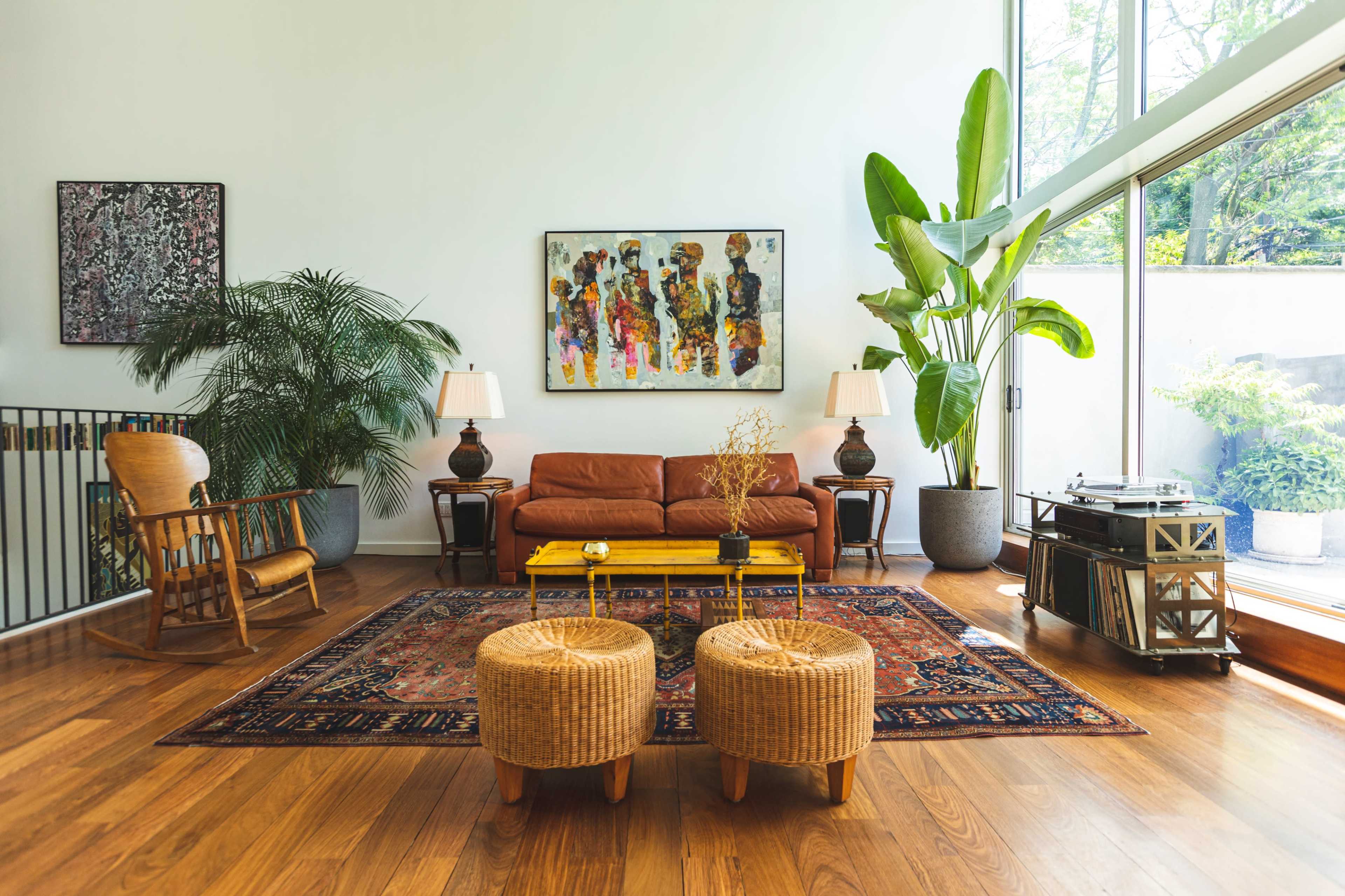 The living room features a brown leather sofa, two wicker stools, a large rug, and a collection of plants, with large windows allowing natural light to fill the space.