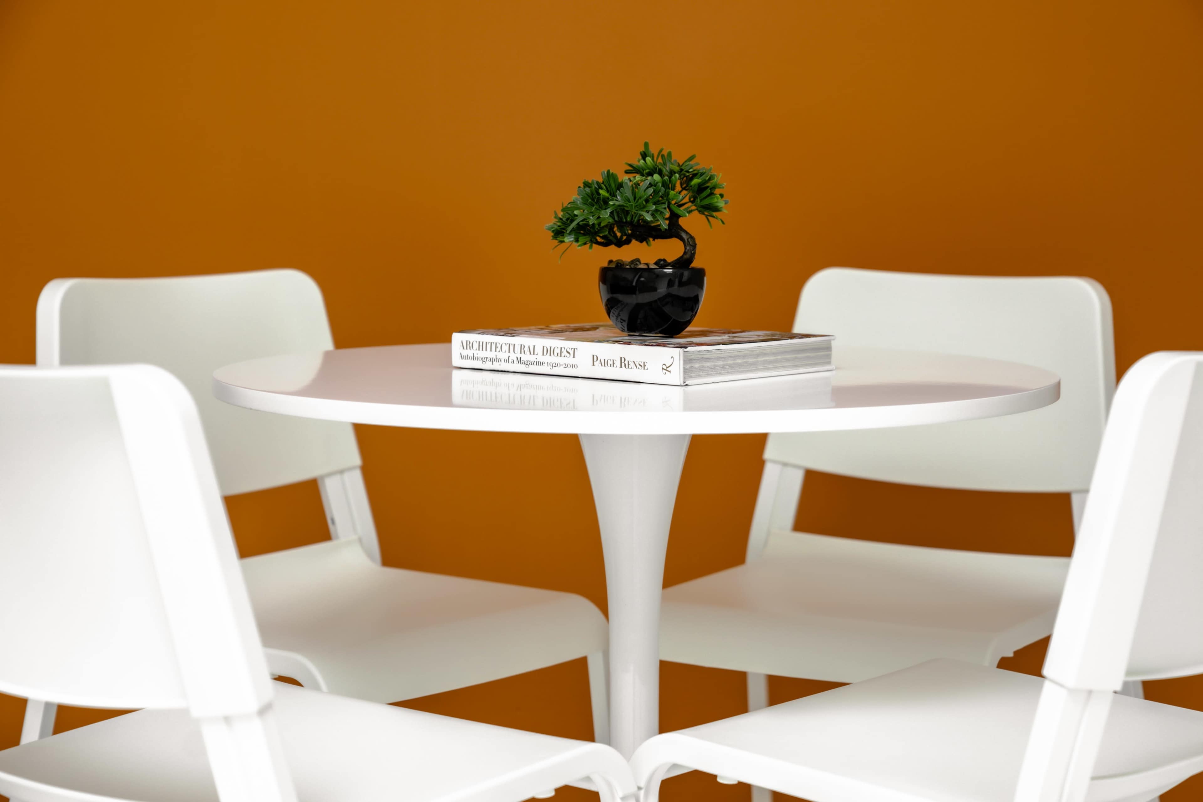 A round white table is surrounded by four white chairs, with a small potted plant and a stack of books on top.