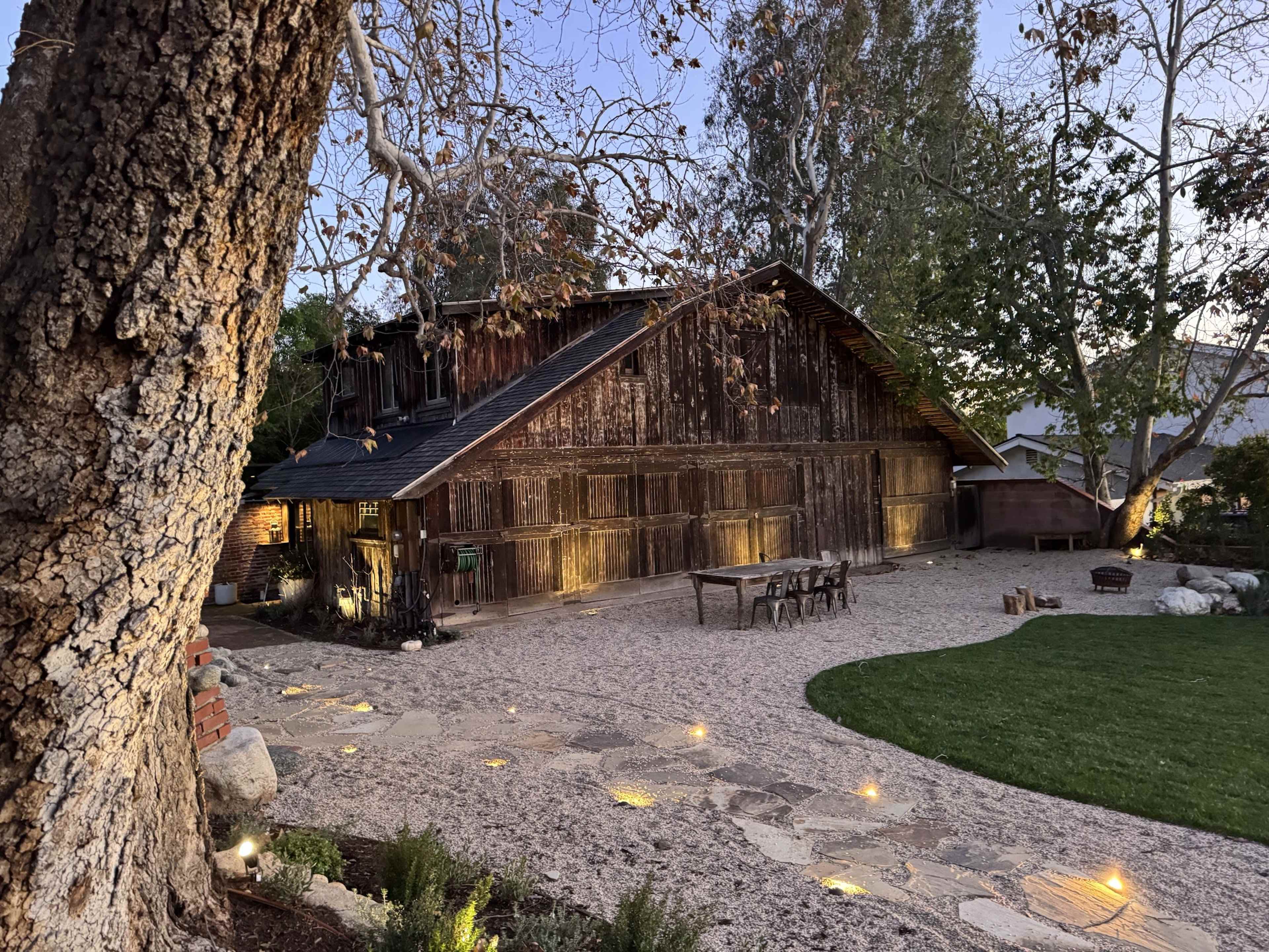 A rustic barn with a weathered wooden exterior is set against a landscaped yard featuring stone pathways and scattered lighting.