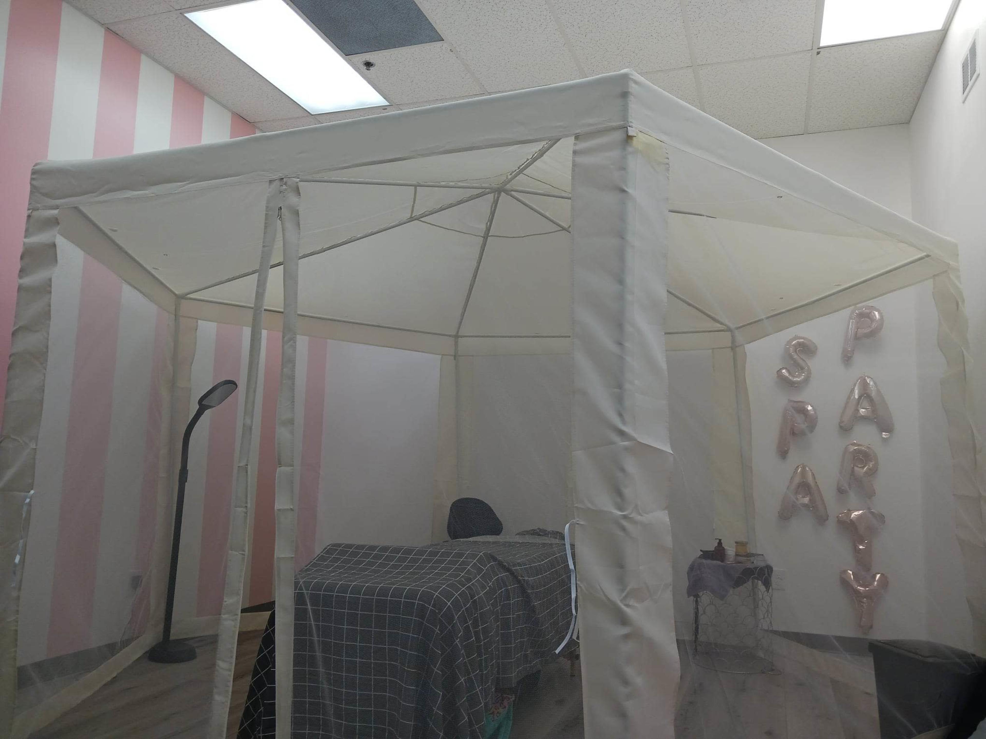 A white canopy tent with an enclosed area is set up in a room, featuring a massage table and decorative lettering that spells out "SPA PARTY."