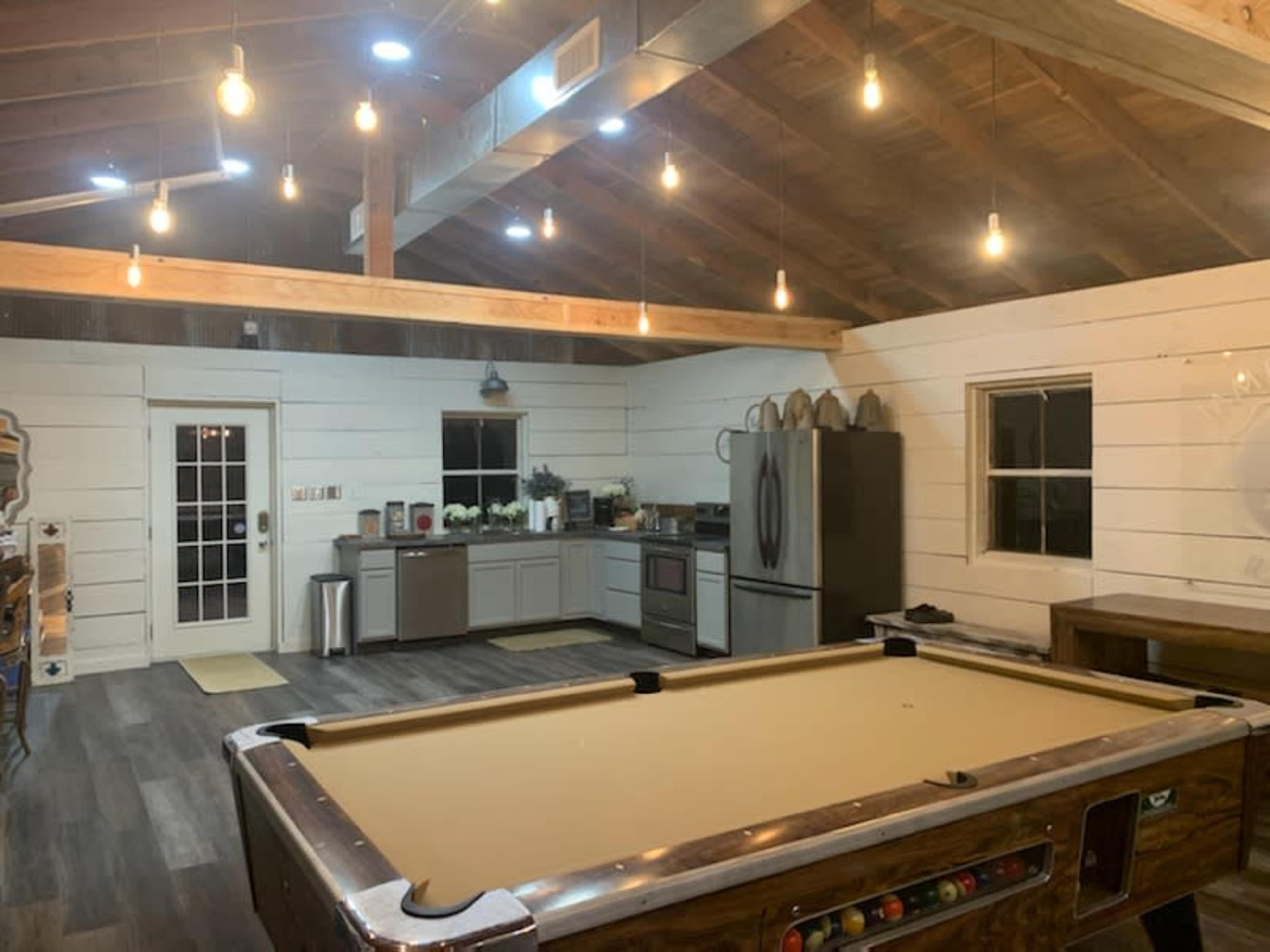A spacious, well-lit room featuring a pool table, a modern kitchen with stainless steel appliances, and white shiplap walls.