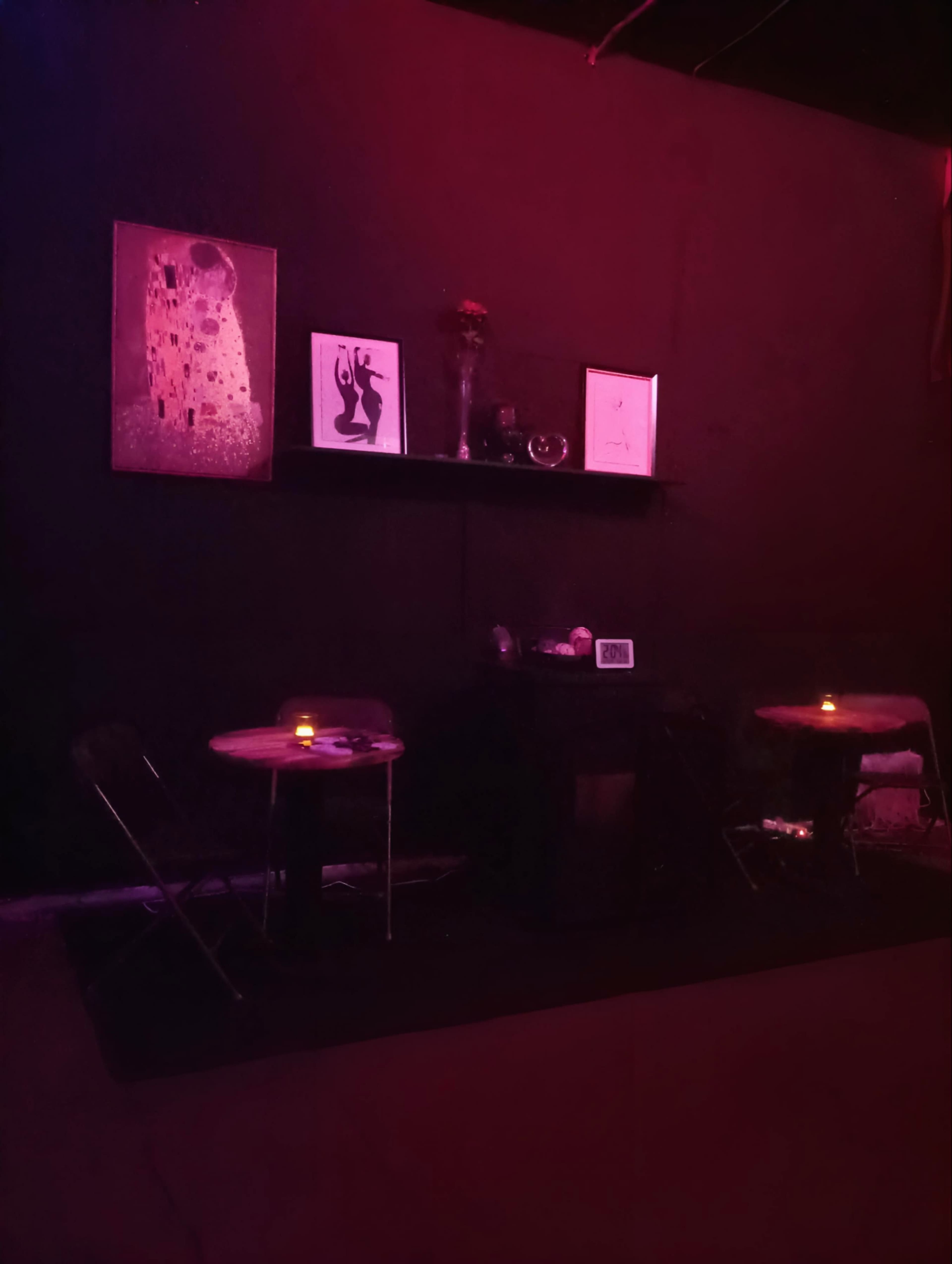 A dimly lit space features two small tables with candles, framed artwork on the walls, and a shelf with decorative items.